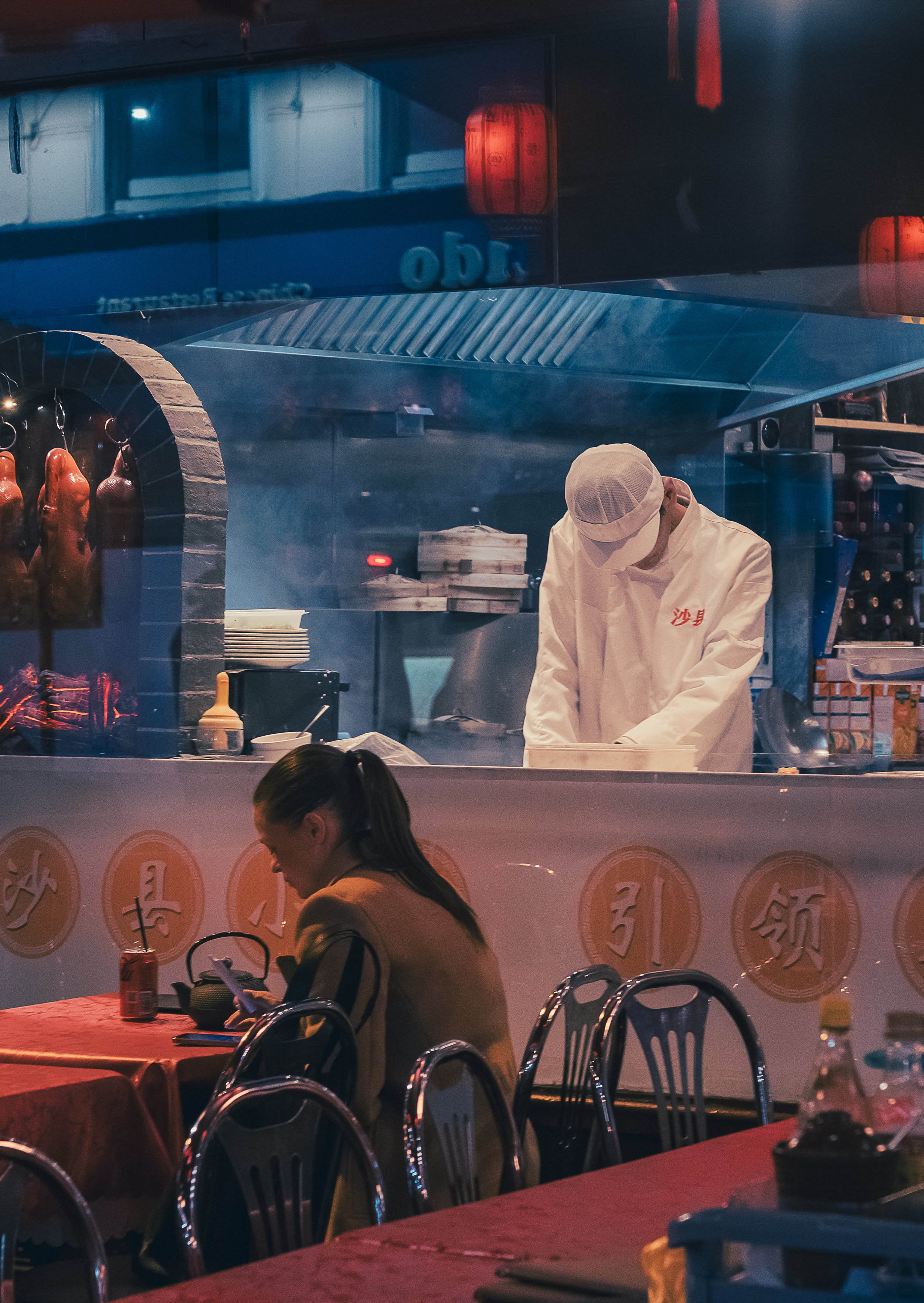 A chef in a white uniform and hat preparing food behind a glass window in a restaurant kitchen, with a woman sitting at a table in the foreground looking at her phone.