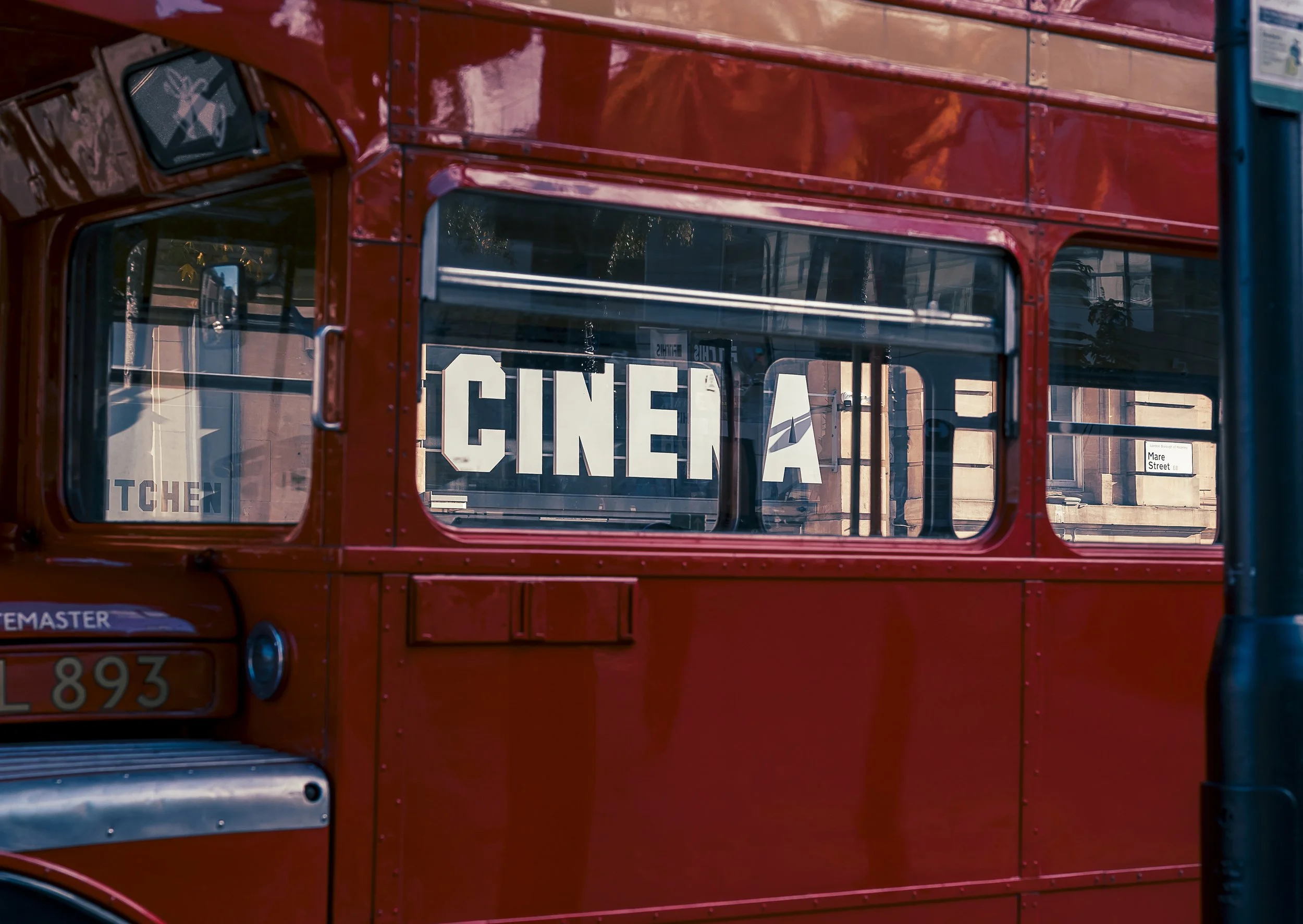 A red double-decker bus with large windows. The word 'CINEMA' is displayed on the side window, and there is a street sign indicating Mare Street in the background.