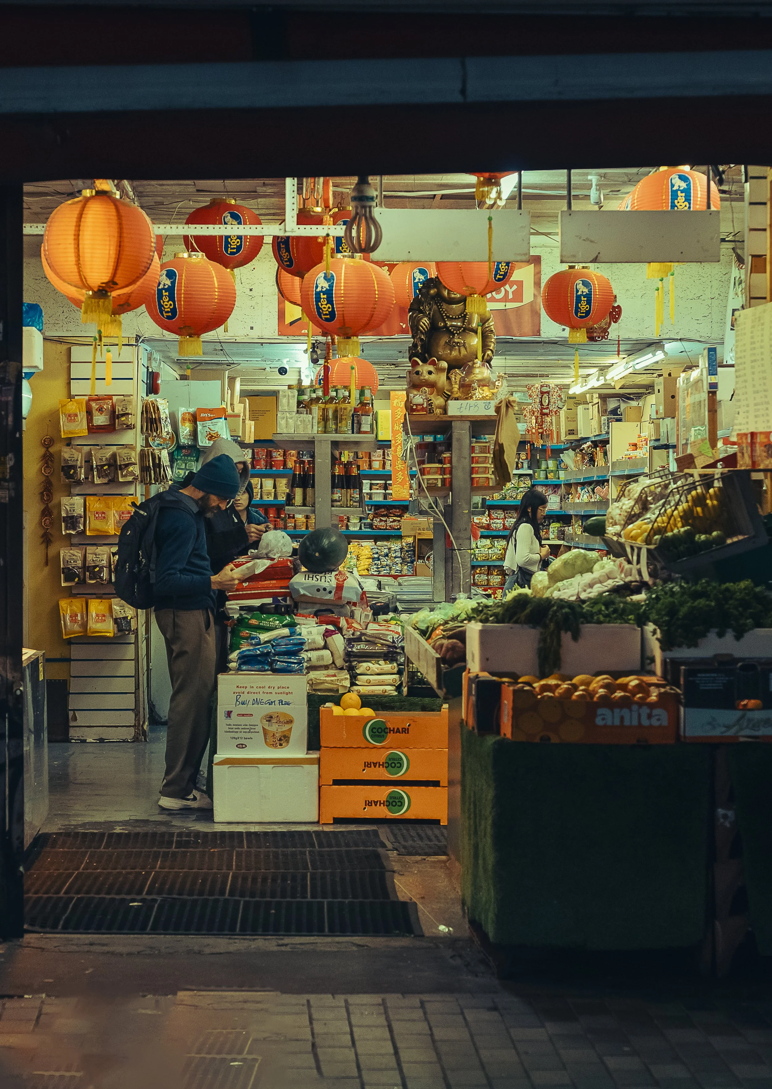 Inside a grocery store with bright overhead lighting, hanging red lanterns, and various food items on shelves and stacked in boxes, including fresh produce.