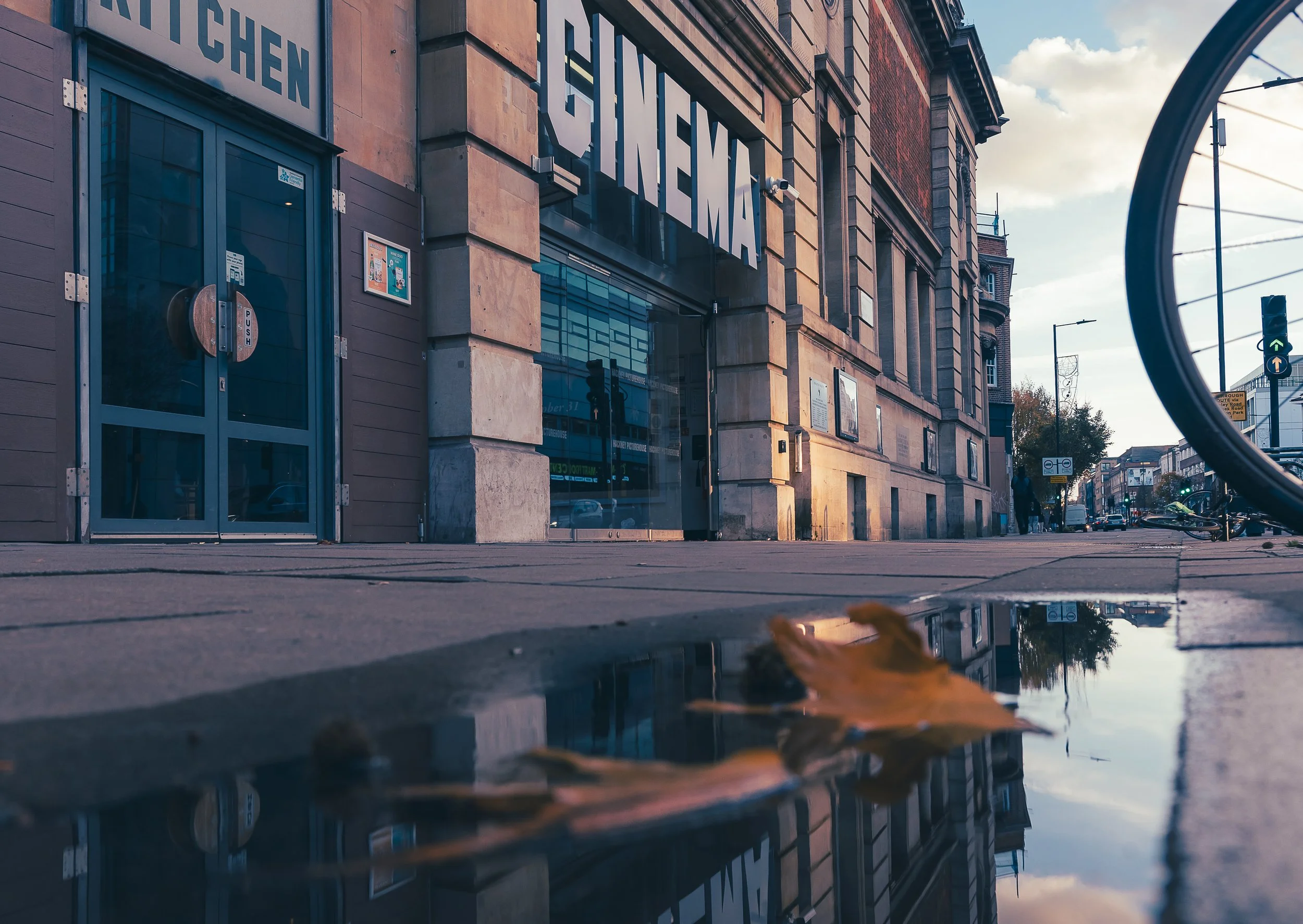 View of a city street with a cinema building, reflecting the sky and surroundings in a puddle in the foreground, with a bicycle partially visible on the right, and traffic lights and pedestrians in the background.