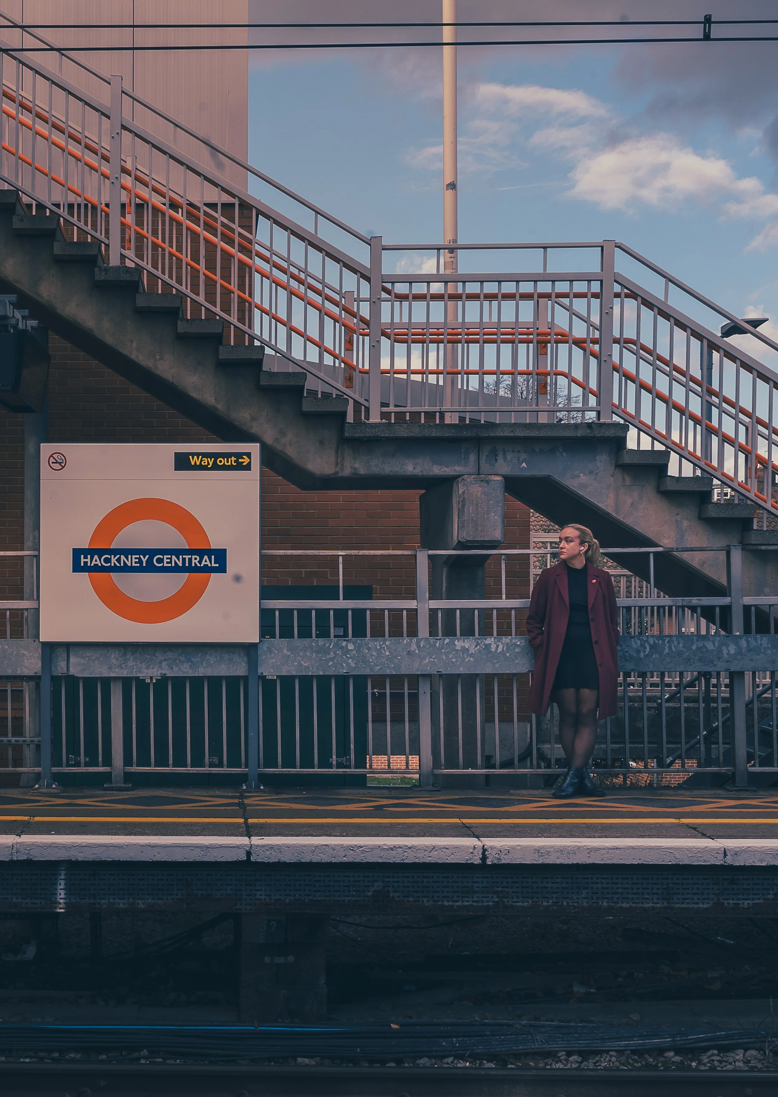 A woman with a maroon coat and black dress standing on a train station platform near a railing, with a sign reading "Hackney Central" and an orange circle logo, under a clear sky with some clouds and a staircase overhead.