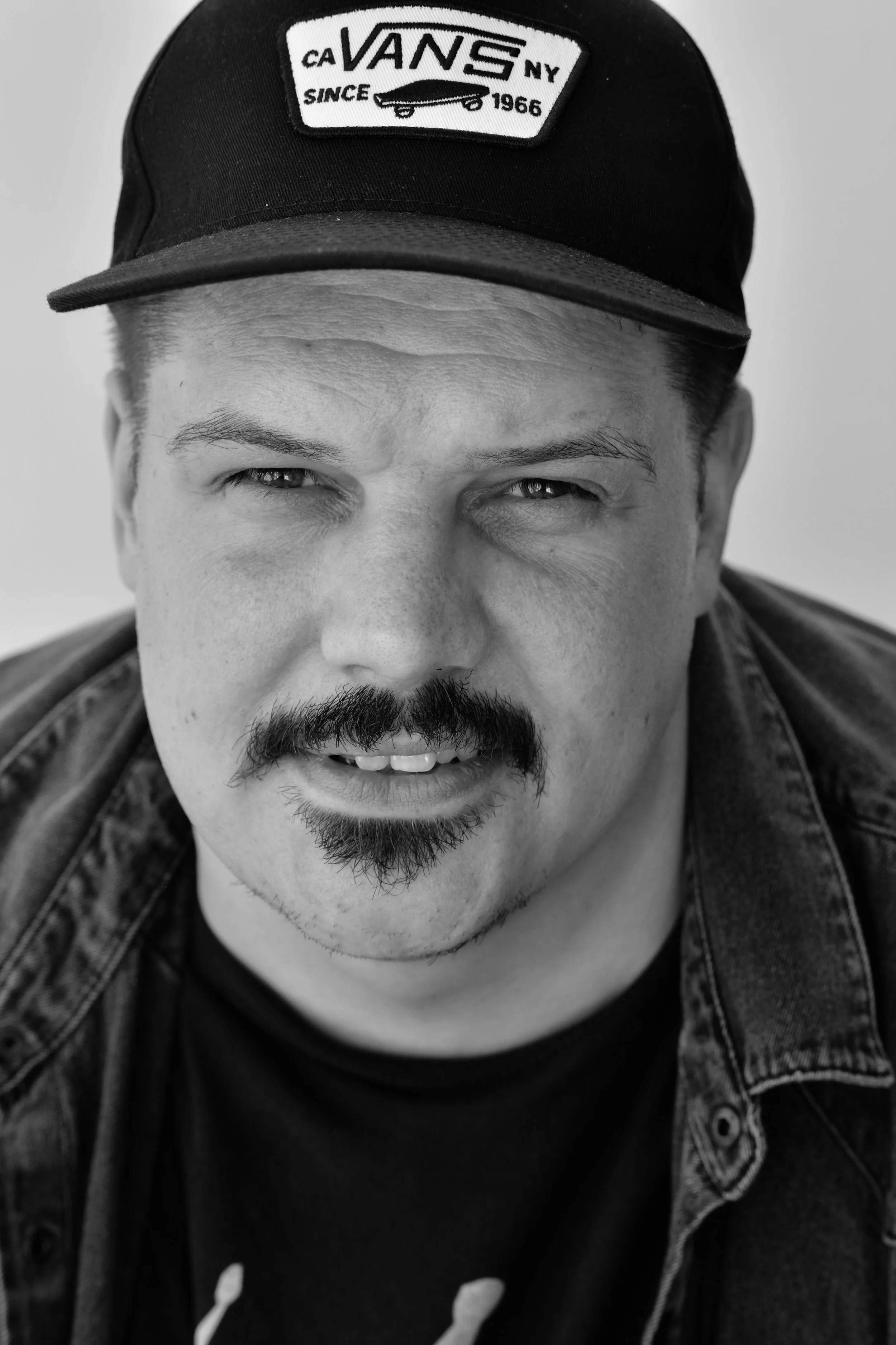 Black and white close-up portrait of a man wearing a Vans cap, a mustache and goatee, with a denim jacket.