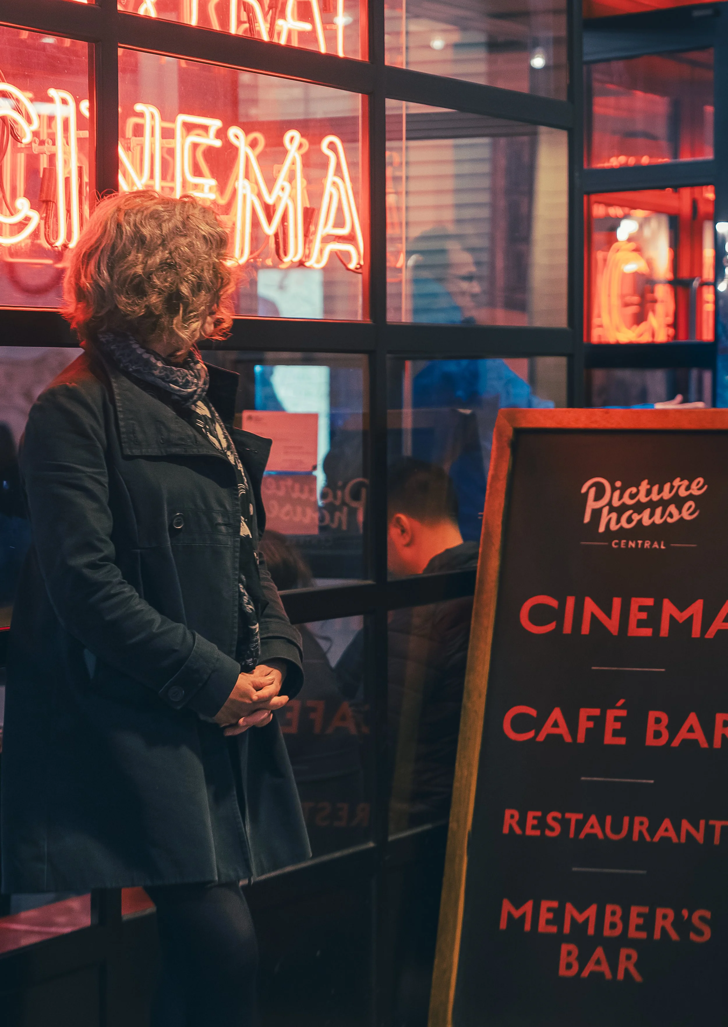 A woman in a black coat and scarf stands outside a cinema, looking in through the window. The window has red neon signs and a blackboard sign lists amenities like cinema, cafe bar, restaurant, and member's bar outside the establishment.
