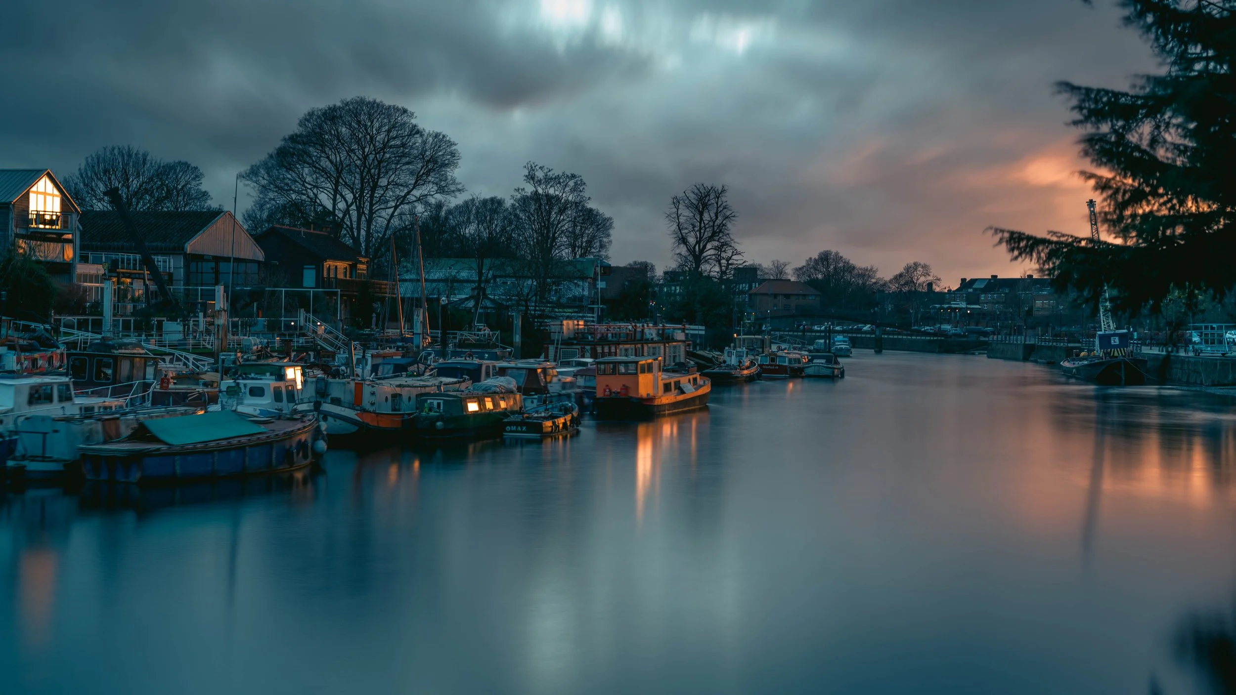 Moored boats along a river at sunset with calm water and cloudy sky.