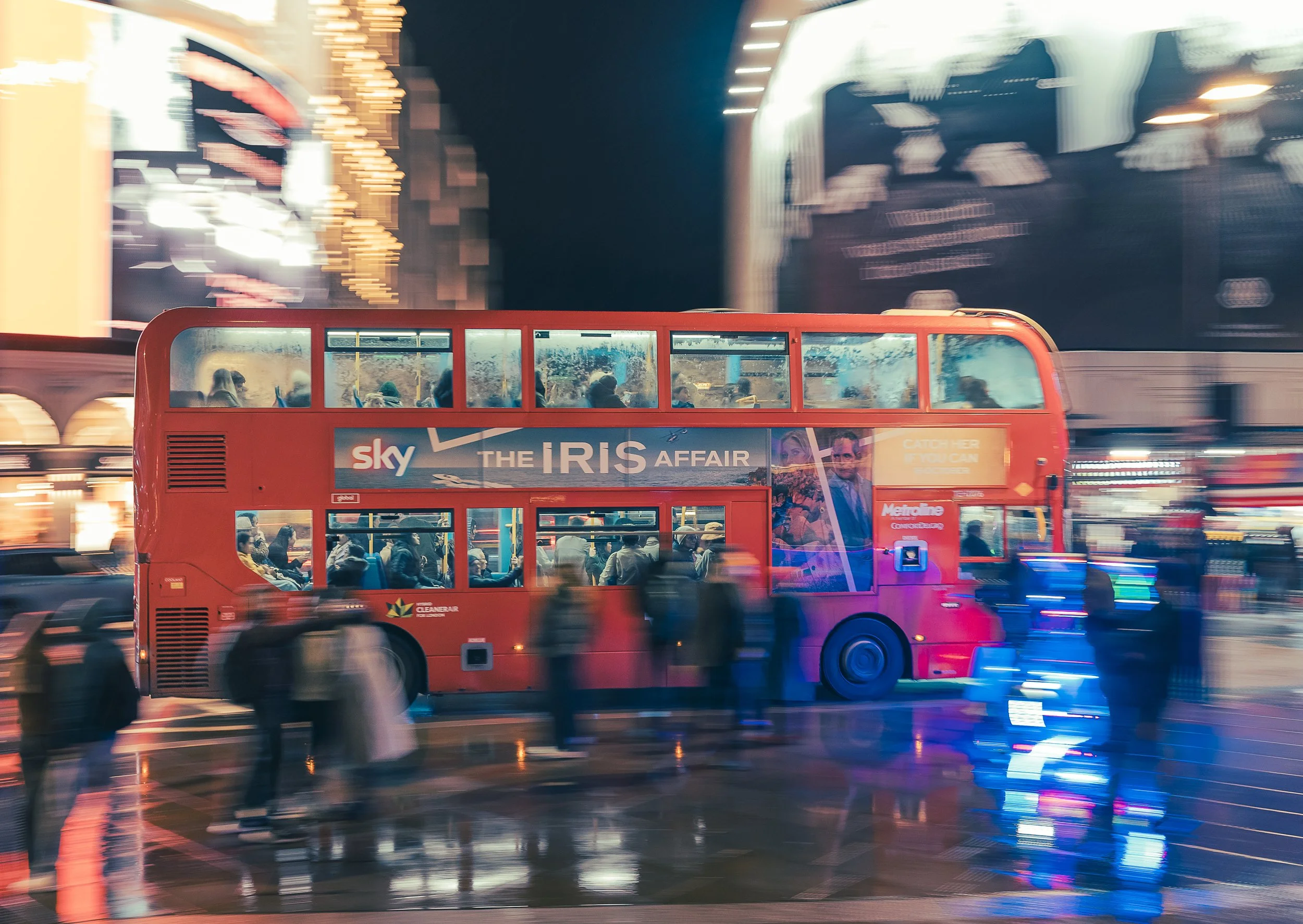 A red double-decker bus with passengers inside and people walking on a city street at night, with bright lights and reflections on wet pavement.