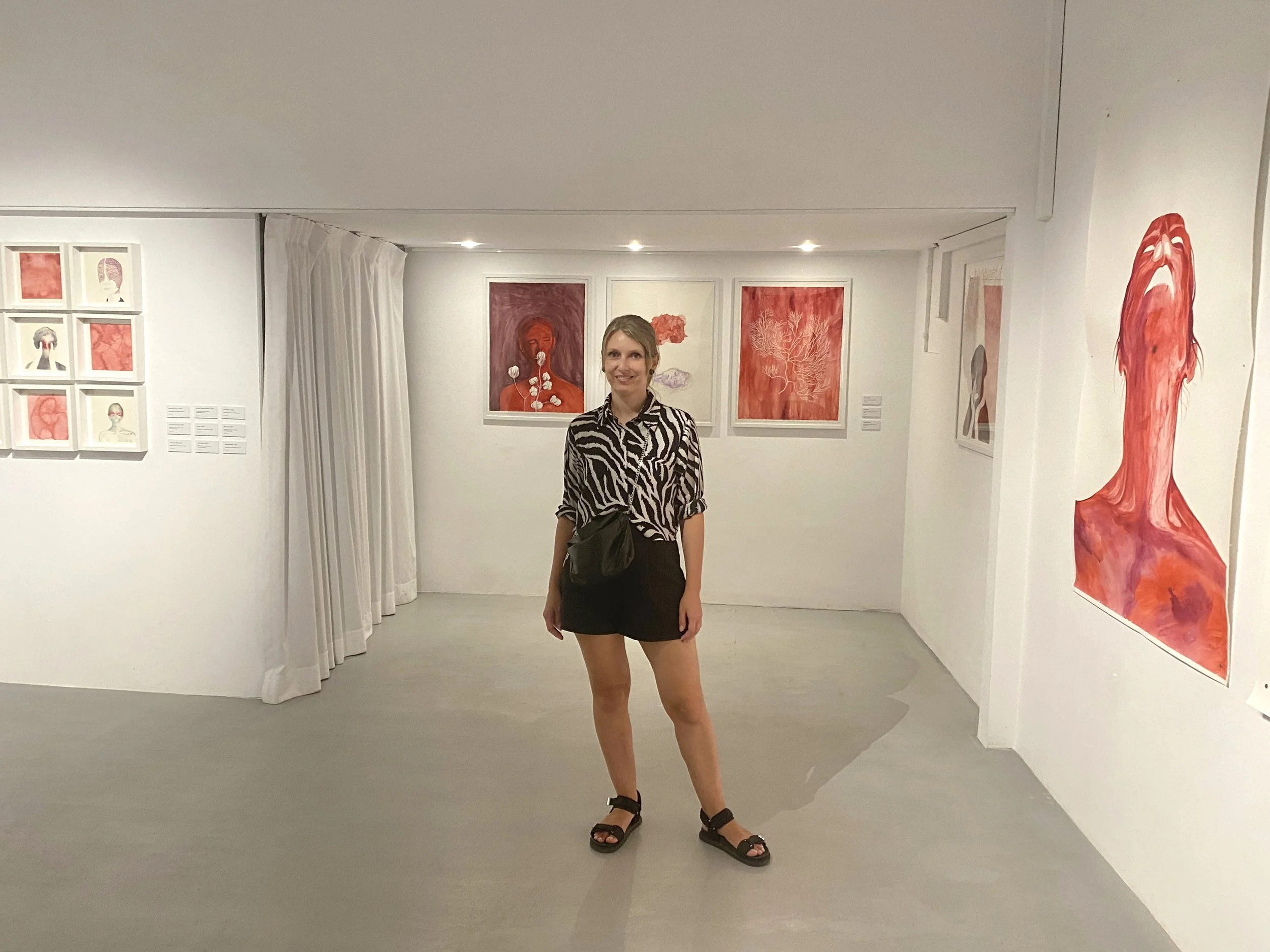 Laura stands in a contemporary art gallery in Bucharest. Behind her are framed paintings featuring watercolor portraits and coral drawings, predominantly in red tones.