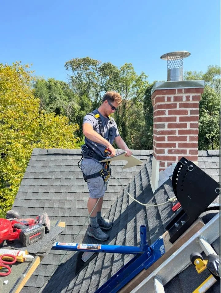 A person working on a roof with tools nearby and a rebuilt and relined brick chimney, surrounded by trees and a clear blue sky.