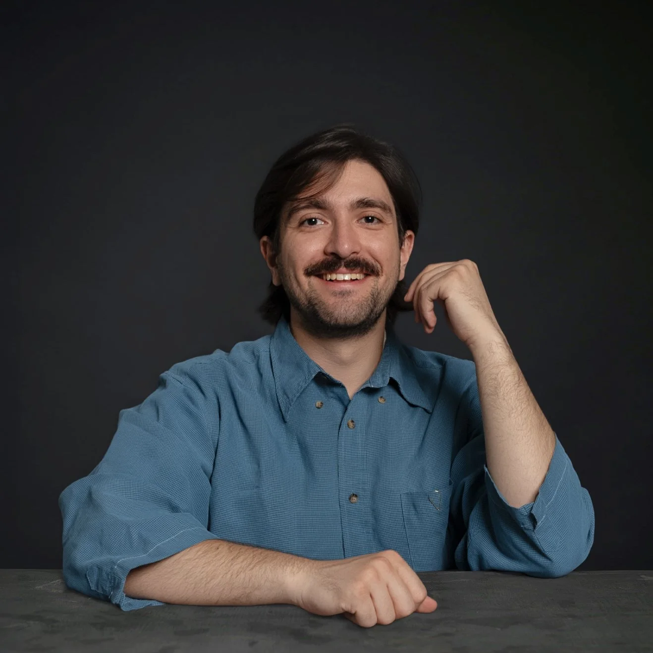 A smiling man with dark hair and a beard wearing a blue button-up shirt, resting his chin on his right hand with his left arm on a dark table against a black background.