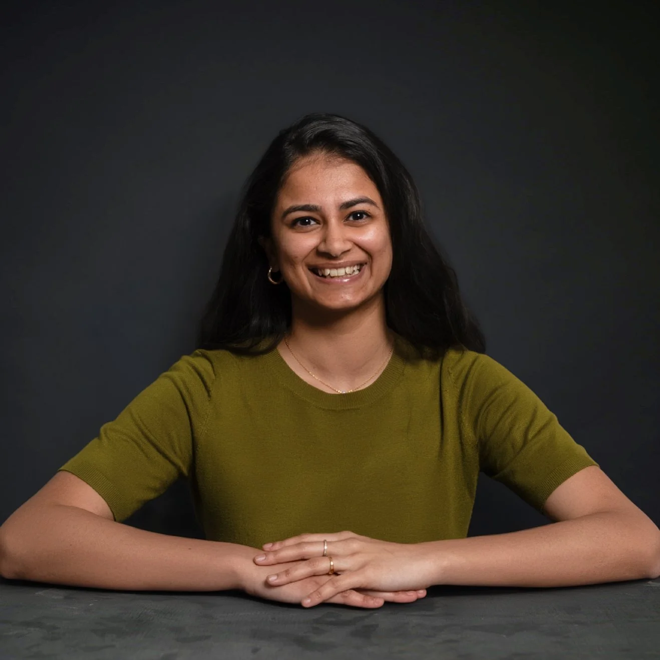 A smiling woman with long dark hair wearing a green short-sleeved shirt, sitting at a table with her arms crossed, against a dark background.