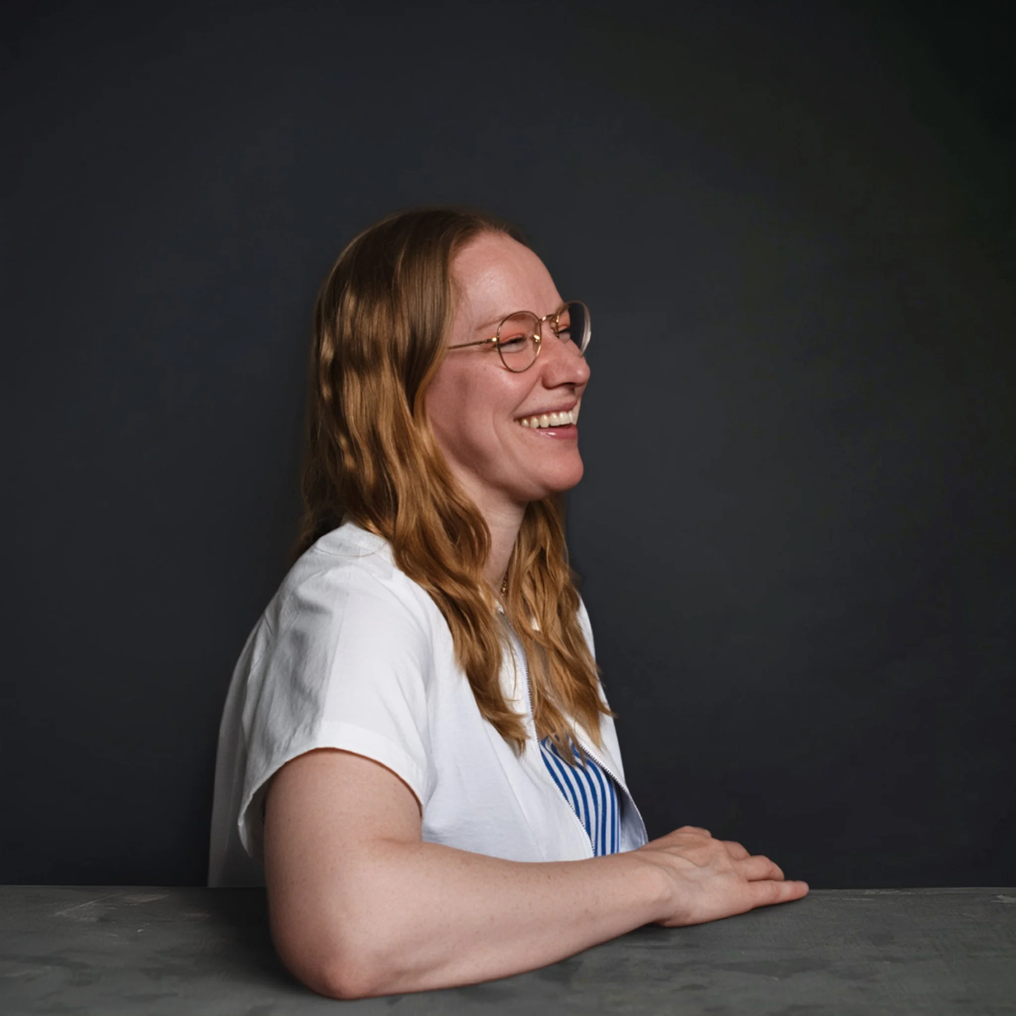 A woman with red hair, glasses, and a white shirt with blue stripes is smiling and resting her head on her folded arms on a dark table against a dark background.
