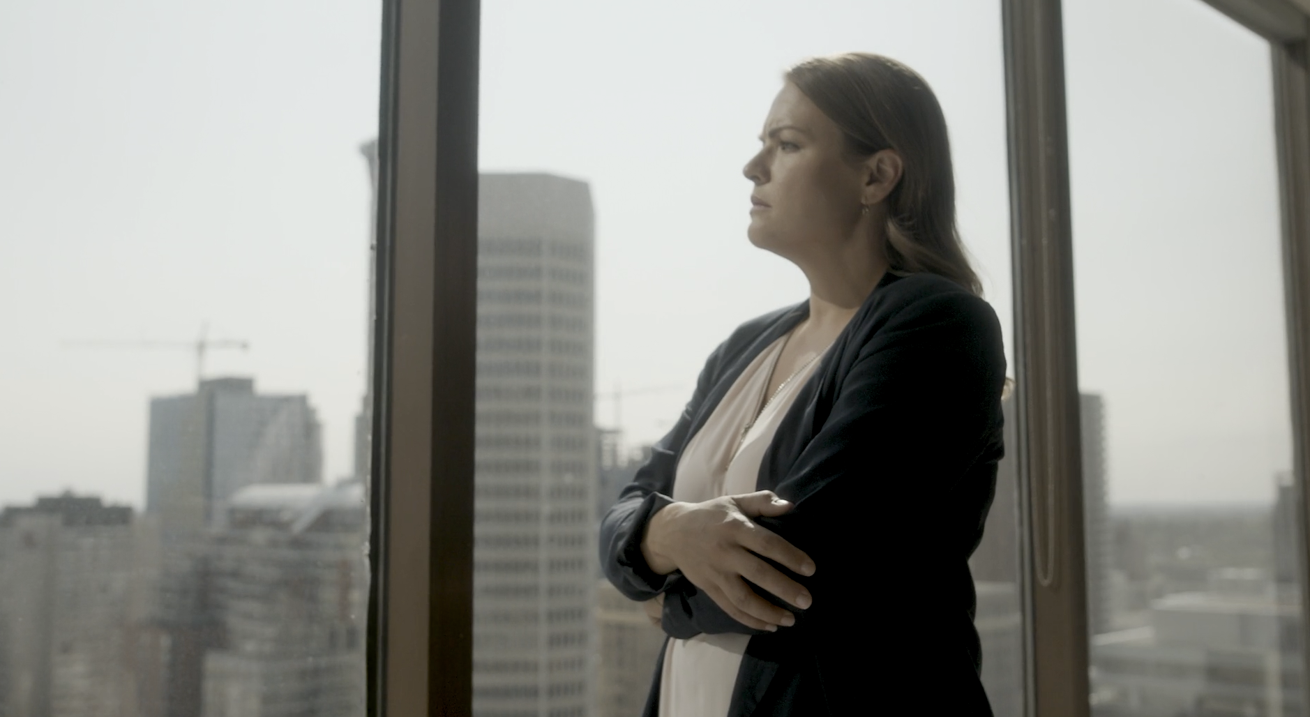 A woman in professional attire standing in front of a large window in a high-rise building, with a city skyline in the background.