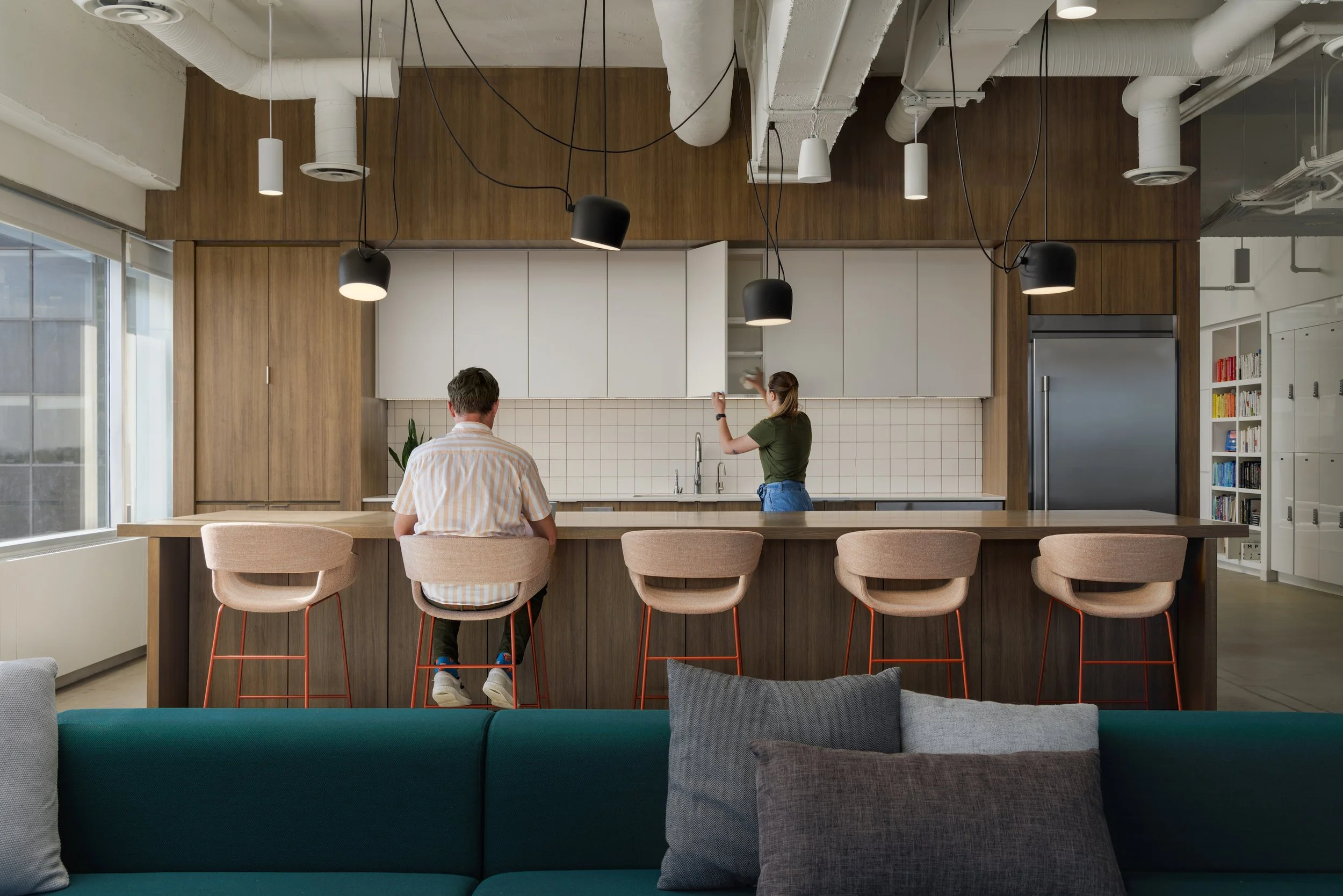Modern open-concept kitchen with white cabinets, wooden accents, and pendant lighting. Two people are standing and sitting at the kitchen island, one woman reaching towards a cabinet. Barstools are lined up at the island, with a sofa and cushions visible in the foreground.