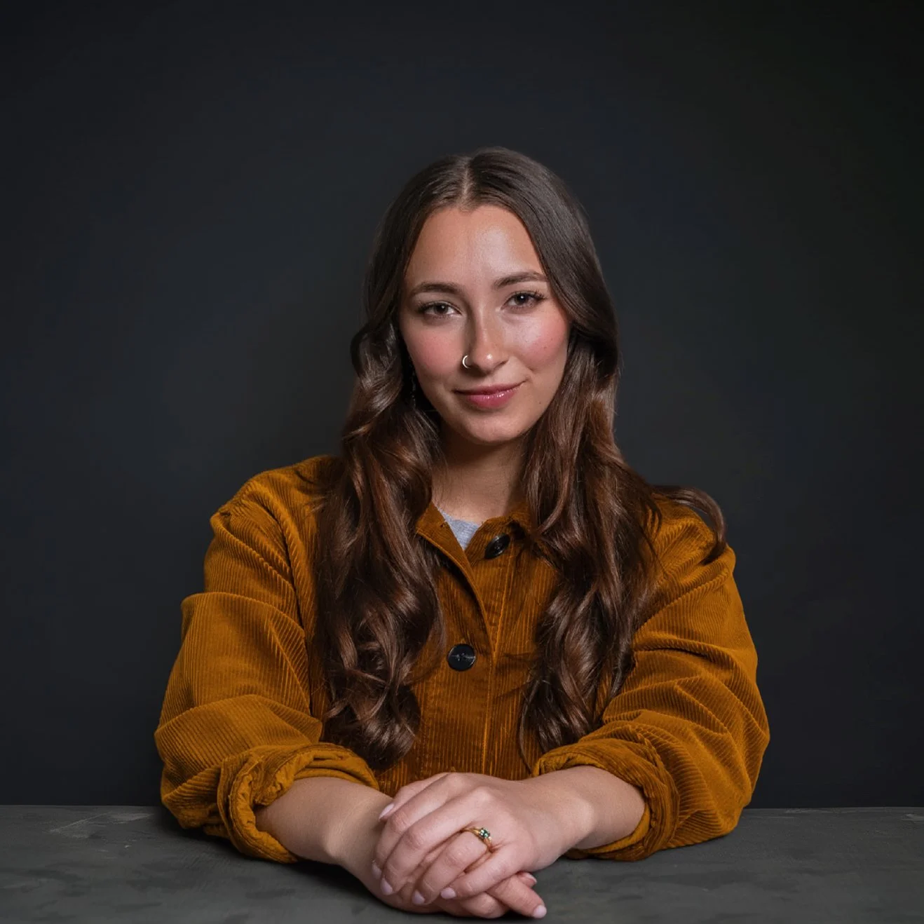 A young woman with long wavy brown hair and a nose ring sitting at a dark table against a plain dark background, wearing a brown corduroy jacket.