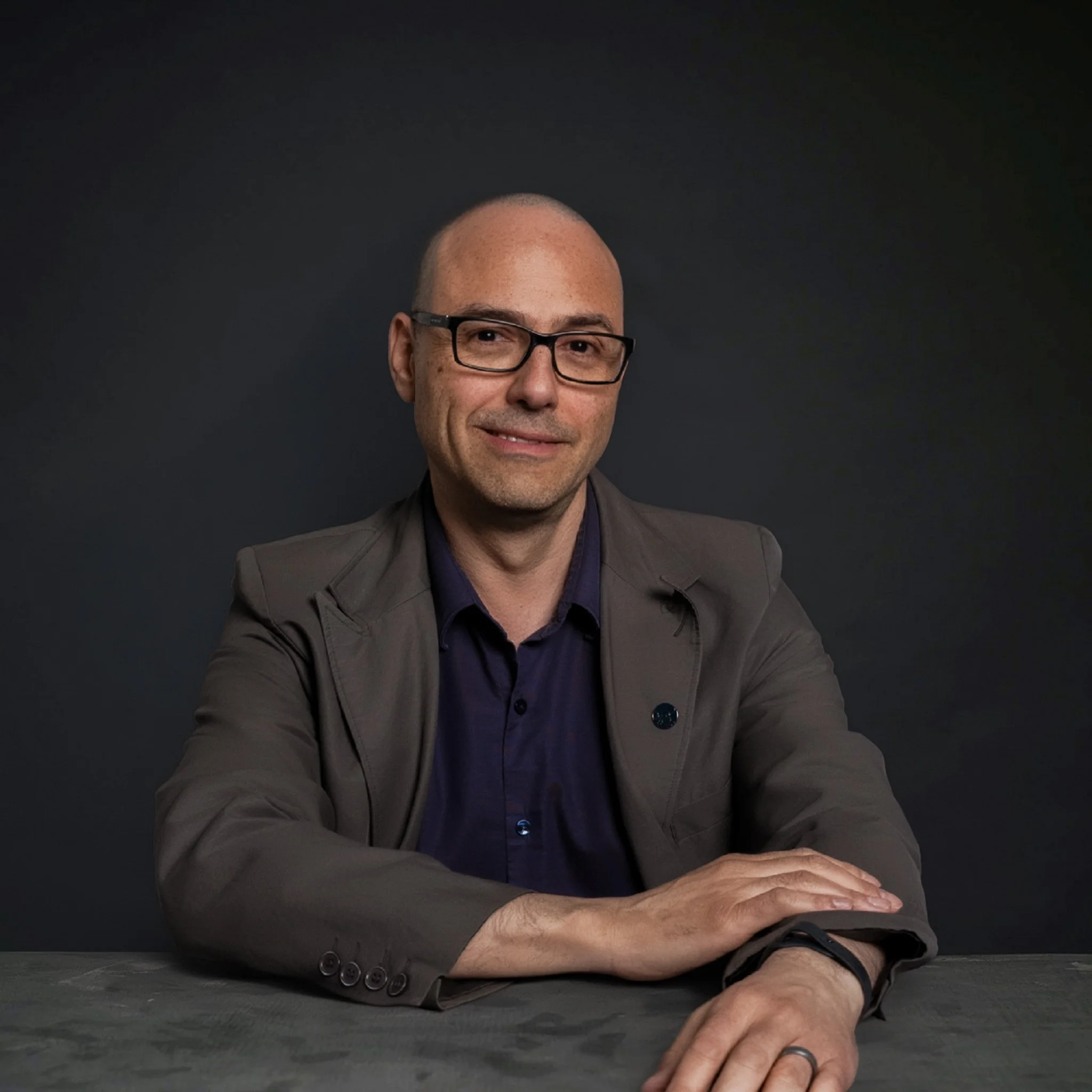 A bald man wearing black glasses and a dark-colored shirt sitting at a table with his arms crossed on the surface, against a dark background.