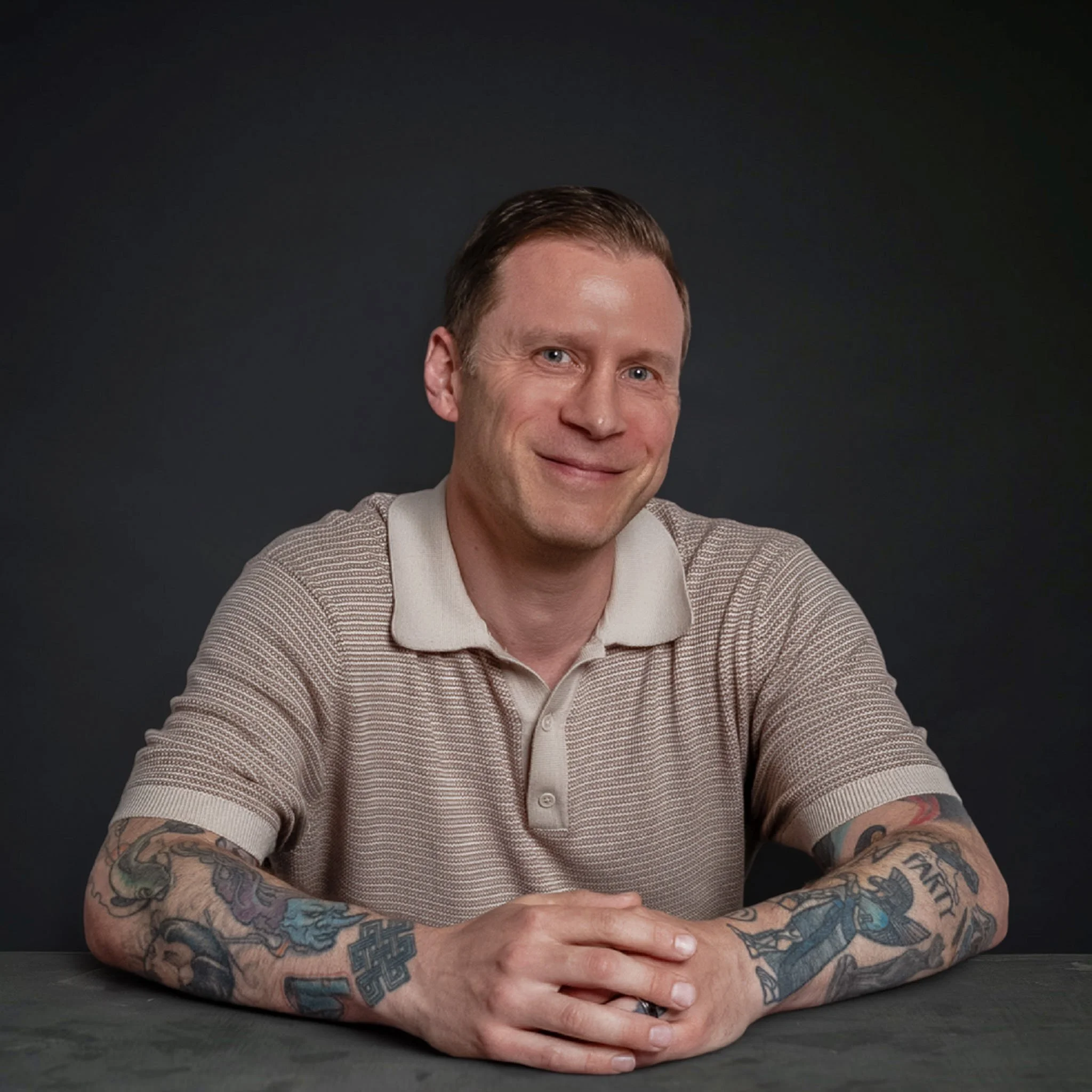 A man with short brown hair and blue eyes smiling, wearing a beige sweater with white collar, is sitting with his arms crossed on a black surface against a dark background.