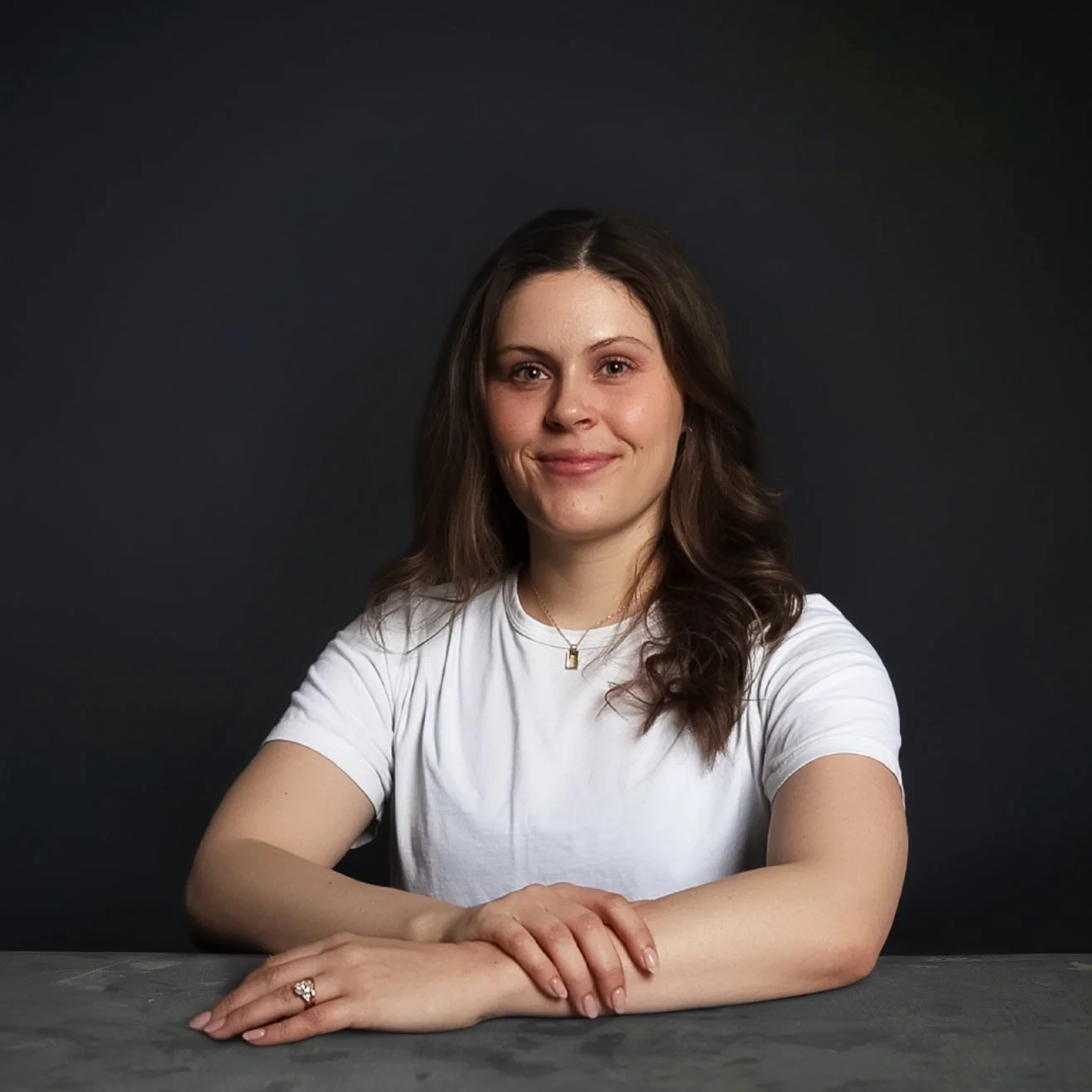 A woman with wavy brown hair wearing a white t-shirt and a necklace, smiling and sitting at a table with her arms crossed, in front of a dark background.