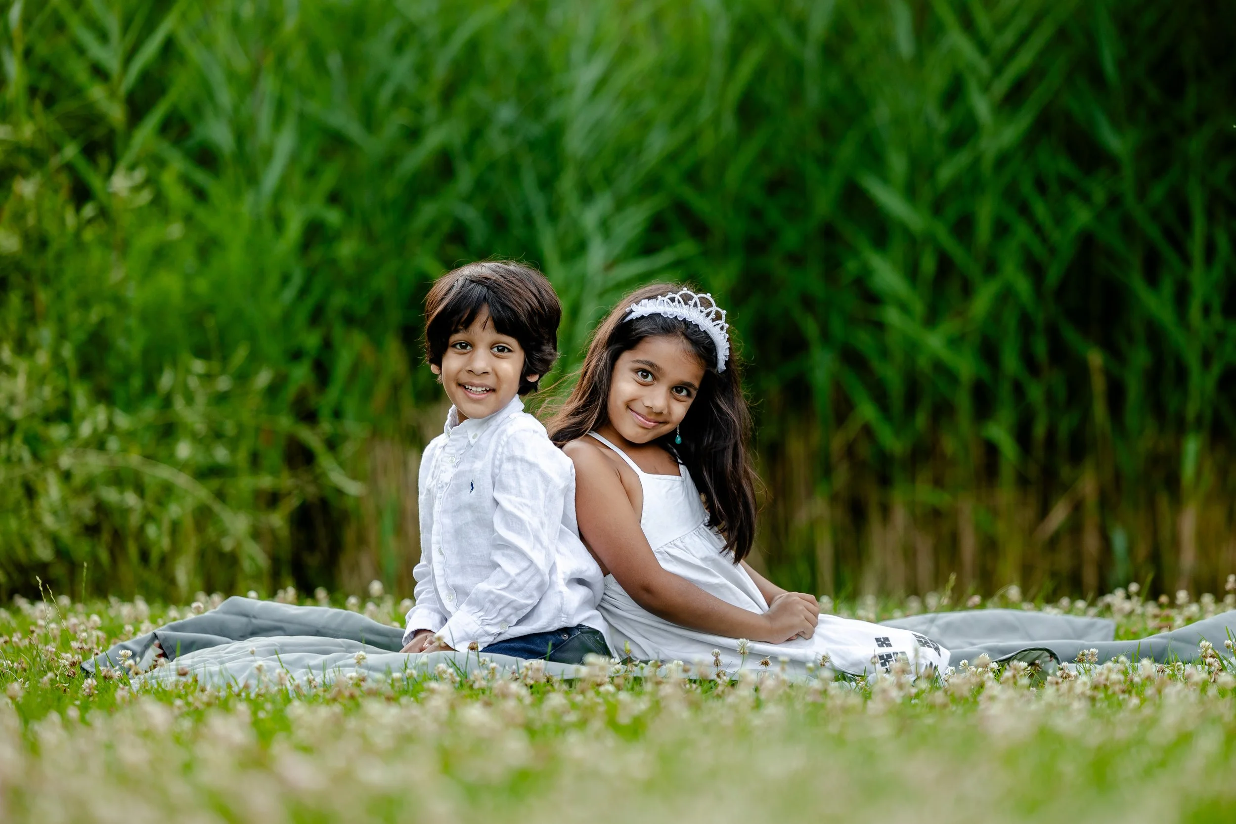 Two children, a boy and a girl, sitting back to back outdoors on a blanket in a field with green plants in the background. The boy has dark, curly hair and wears a white shirt. The girl has long dark hair with a white headband and is wearing a white 