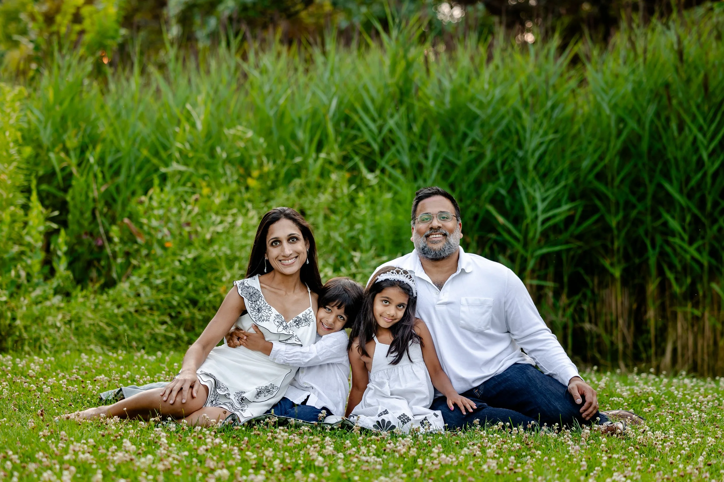 Family of four sitting on grass in front of tall green plants. The mother and father are smiling, with the children sitting between them.