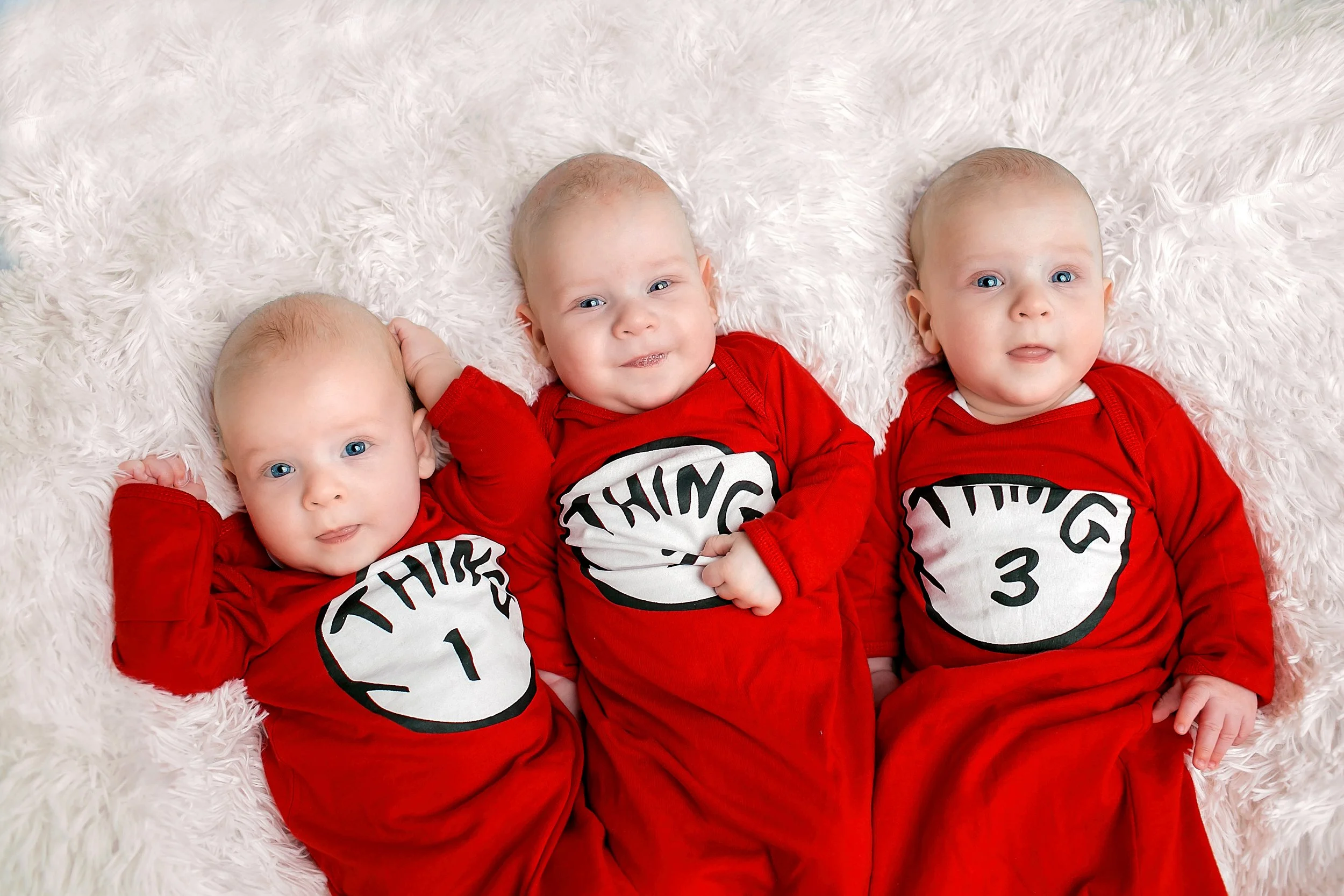 Three babies lying on a fluffy white surface, wearing matching red pajamas with a white clock face and the words "Thing 1," "Thing 2," and "Thing 3" on them. The babies are looking toward the camera with curious and playful expressions.