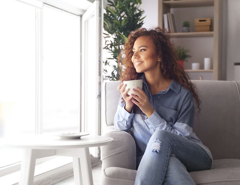 A young woman with curly hair sitting on a sofa, holding a mug and smiling, in a bright living room with large windows and indoor plants.