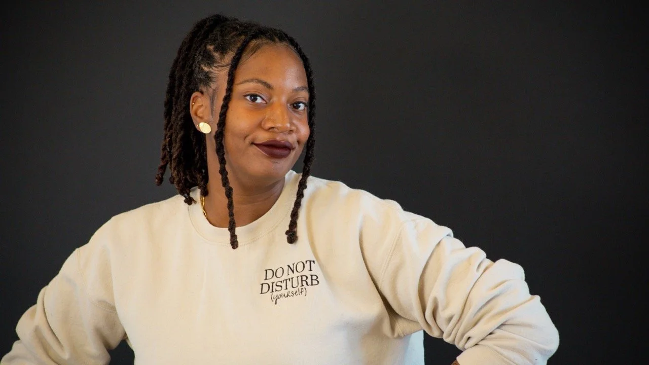 A woman with dreadlocks and dark lipstick posing against a black background, wearing a cream-colored sweatshirt that says "Do Not Disturb (yourself)".