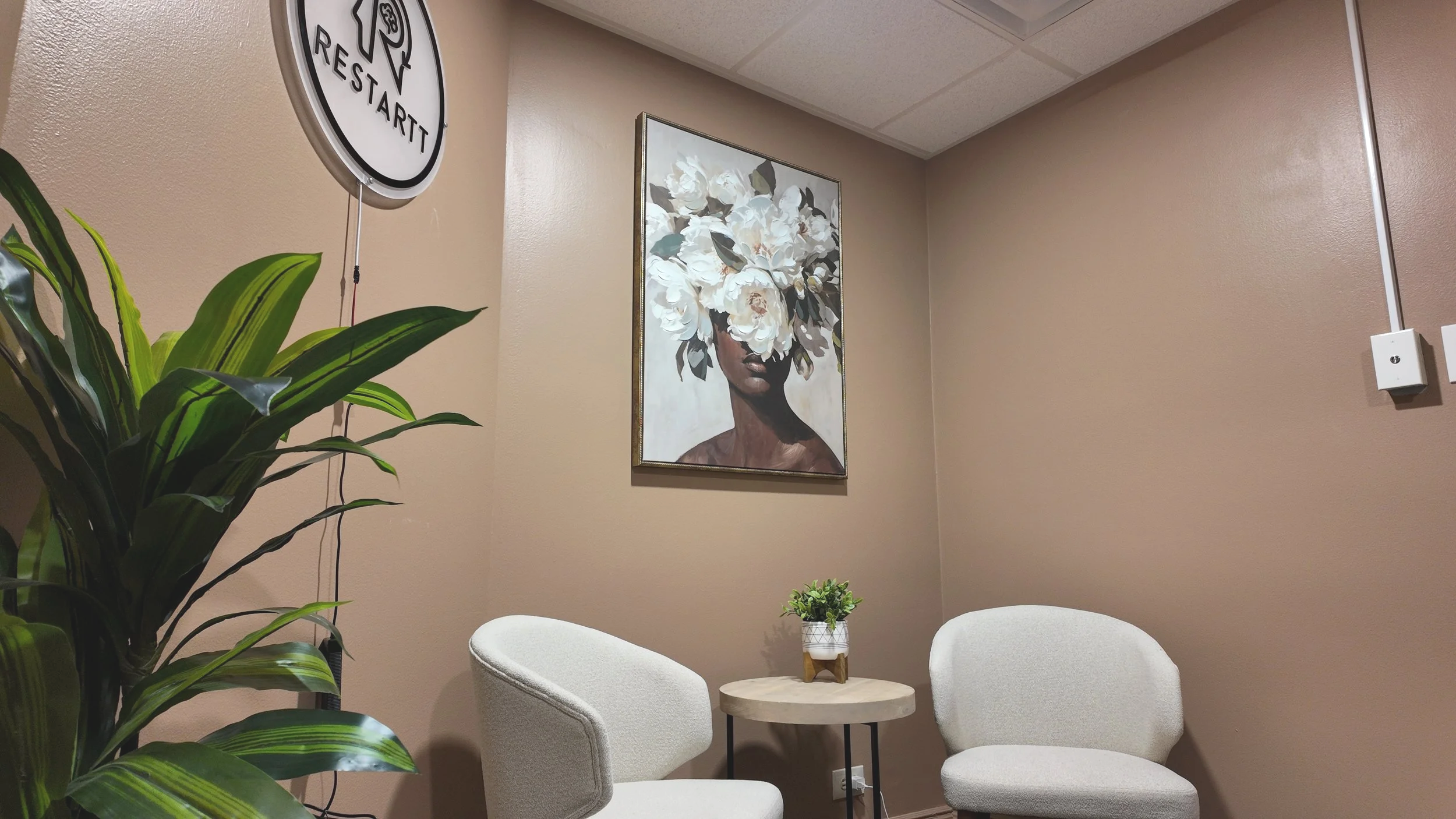 A small seating area with three white chairs and a round wooden table in a room with beige walls. There is a large green plant in the foreground, a small potted plant on the table, and a framed floral portrait on the wall. Part of a wall sign with the text "RESTARTT" is visible.