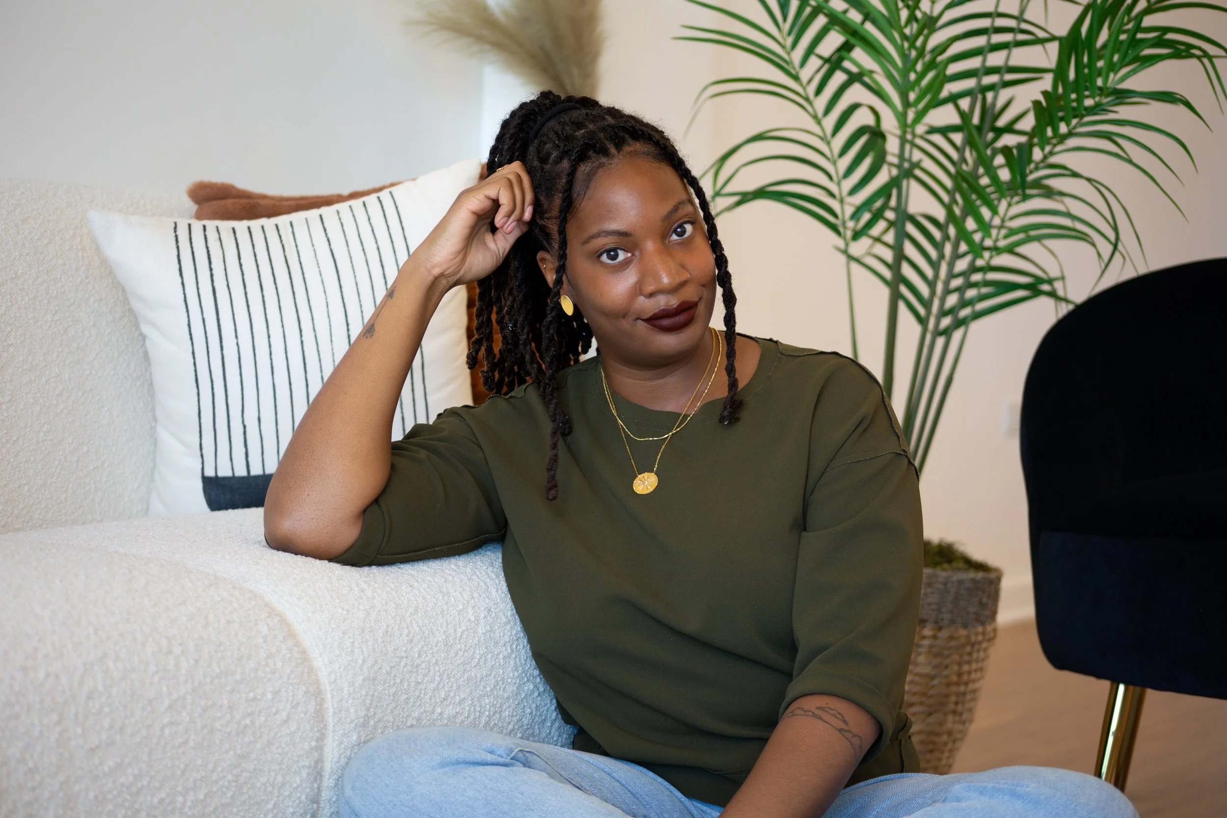 A woman with braided hair and dark lipstick sitting on a white sofa with striped pillows, next to a green potted plant, looking at the camera.