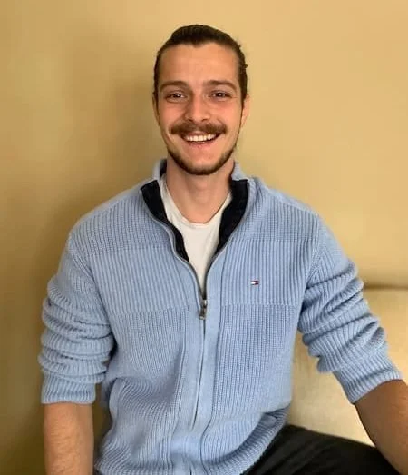 A young man with a beard and mustache, smiling, wearing a light blue zip-up sweater over a white shirt, seated indoors against a beige wall.