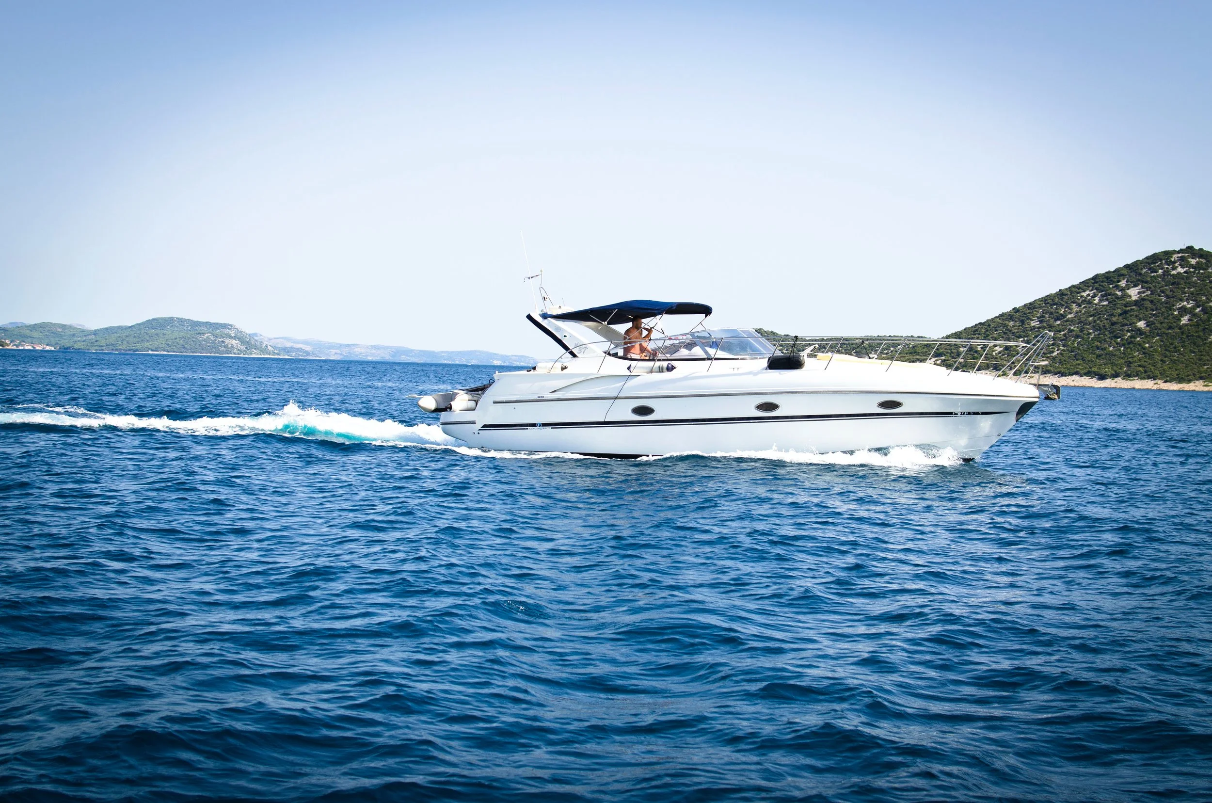 A white motorboat with a black canopy moving through blue water near a mountainous coastline with green foliage.