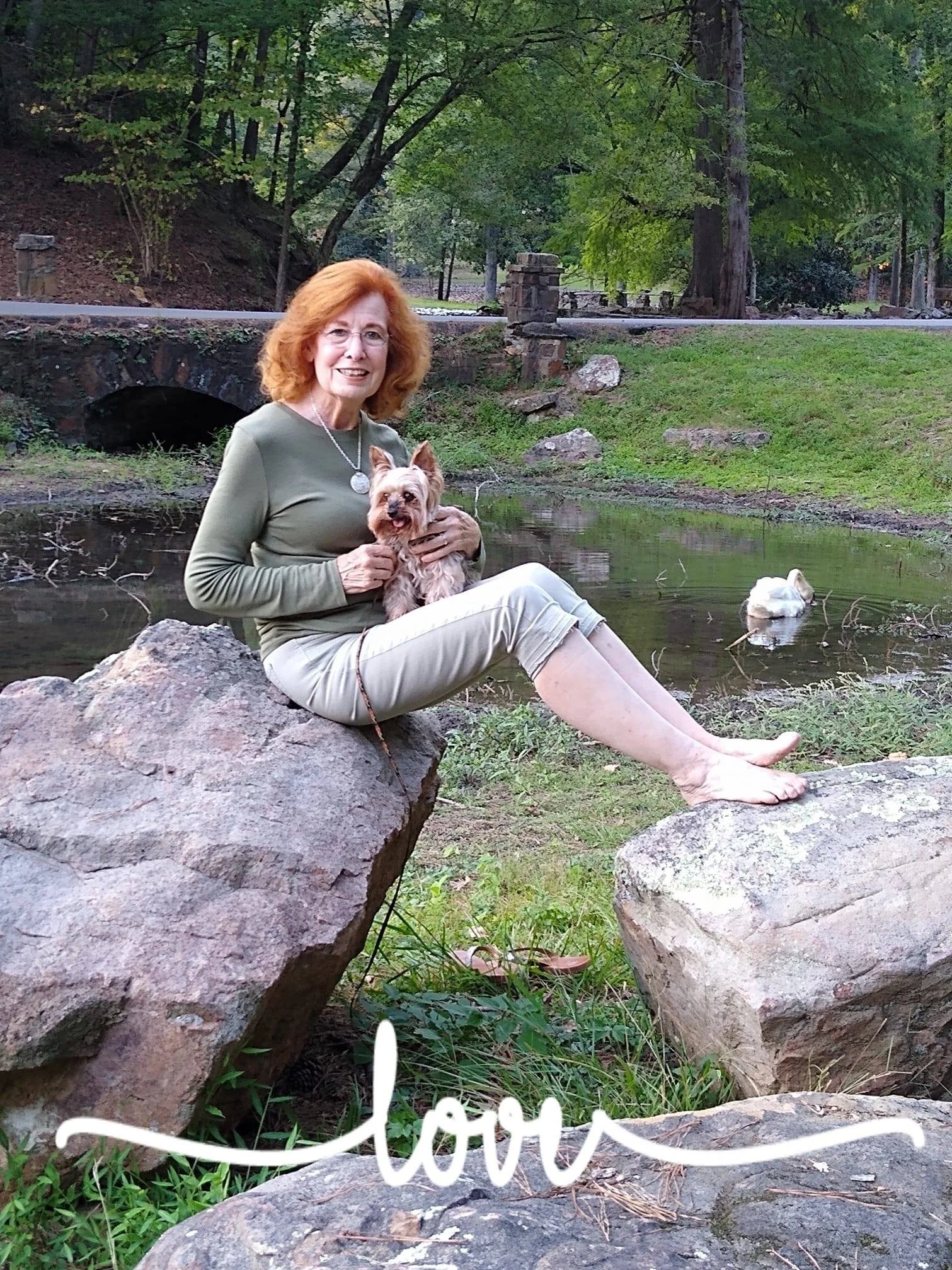 Woman sitting on a rock holding a small dog near water with greenery and a stone bridge in the background, "love" written at the bottom.