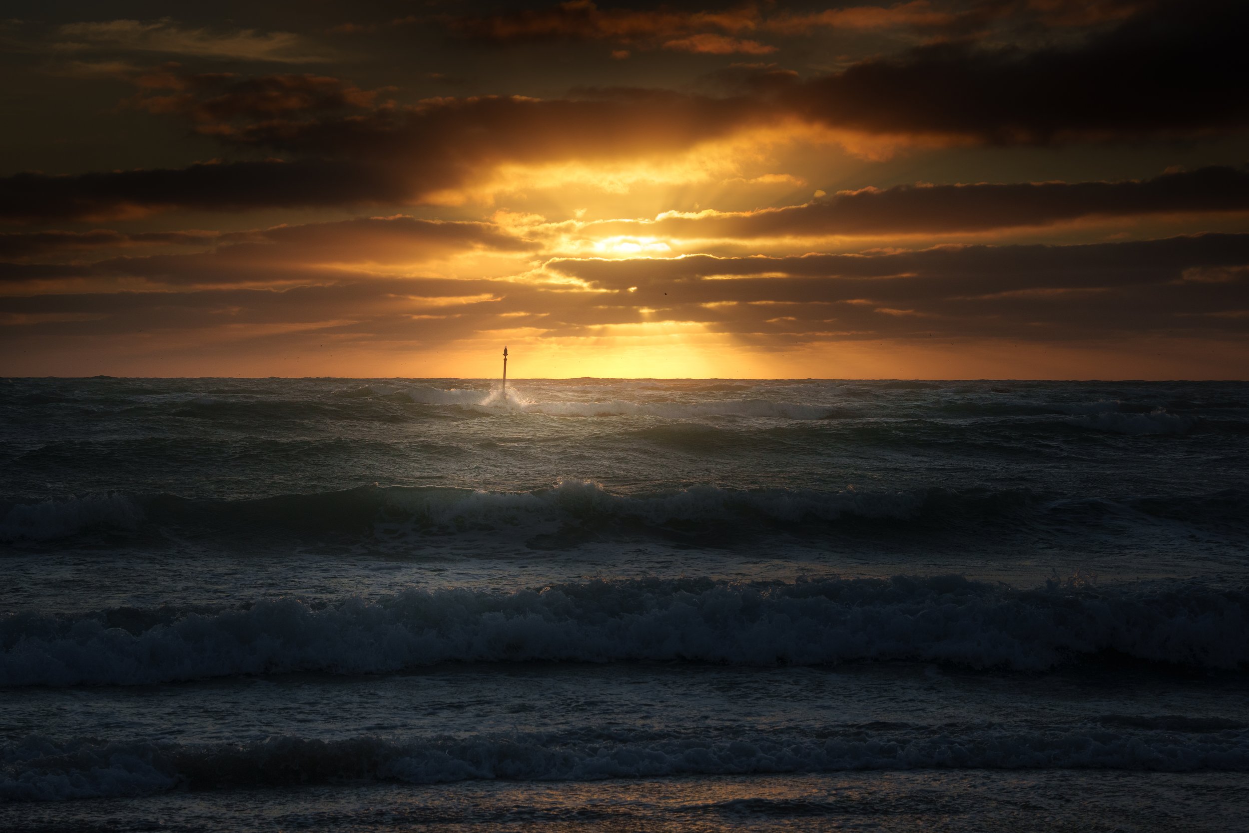 Sunset over the ocean with dark clouds and waves, with a buoy or marker in the distance.