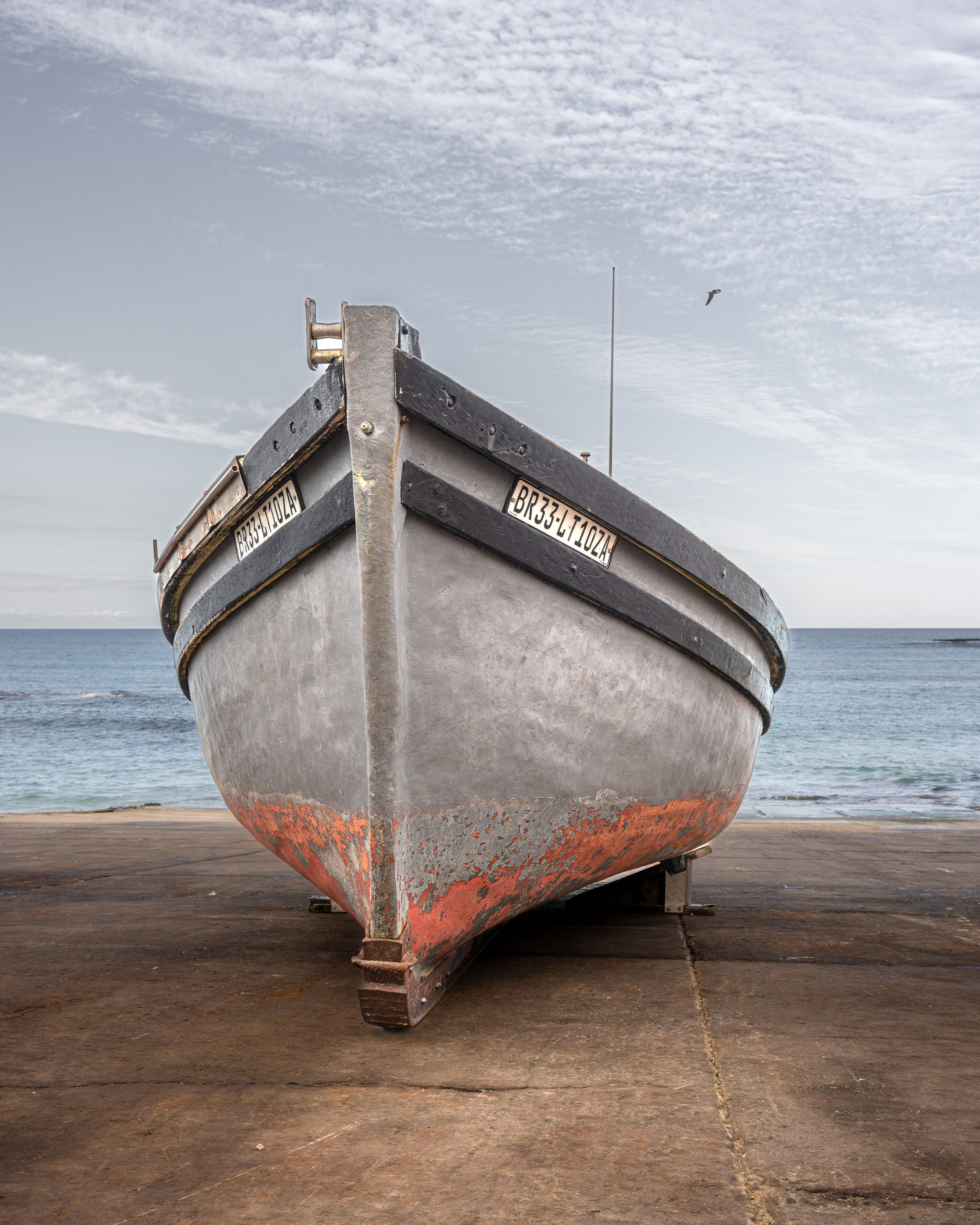 A close-up view of a weathered boat resting on a dock with ocean and cloudy sky in the background.