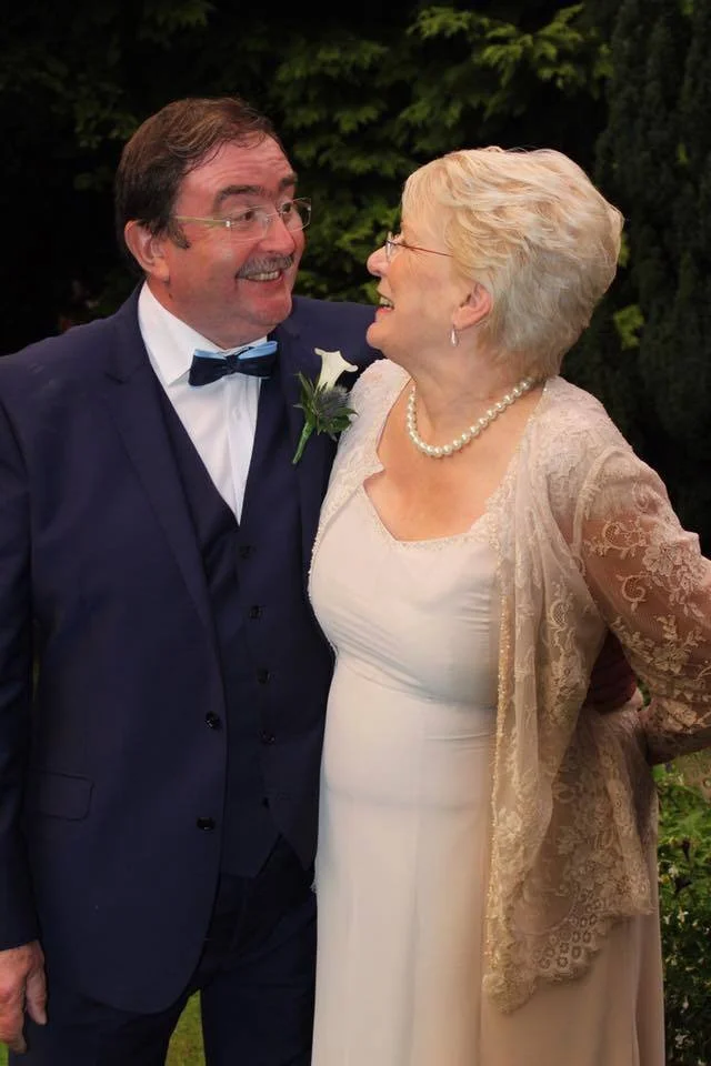 Older couple smiling at each other; man in a suit with bow tie and woman in a lace dress with pearl necklace, outdoors.