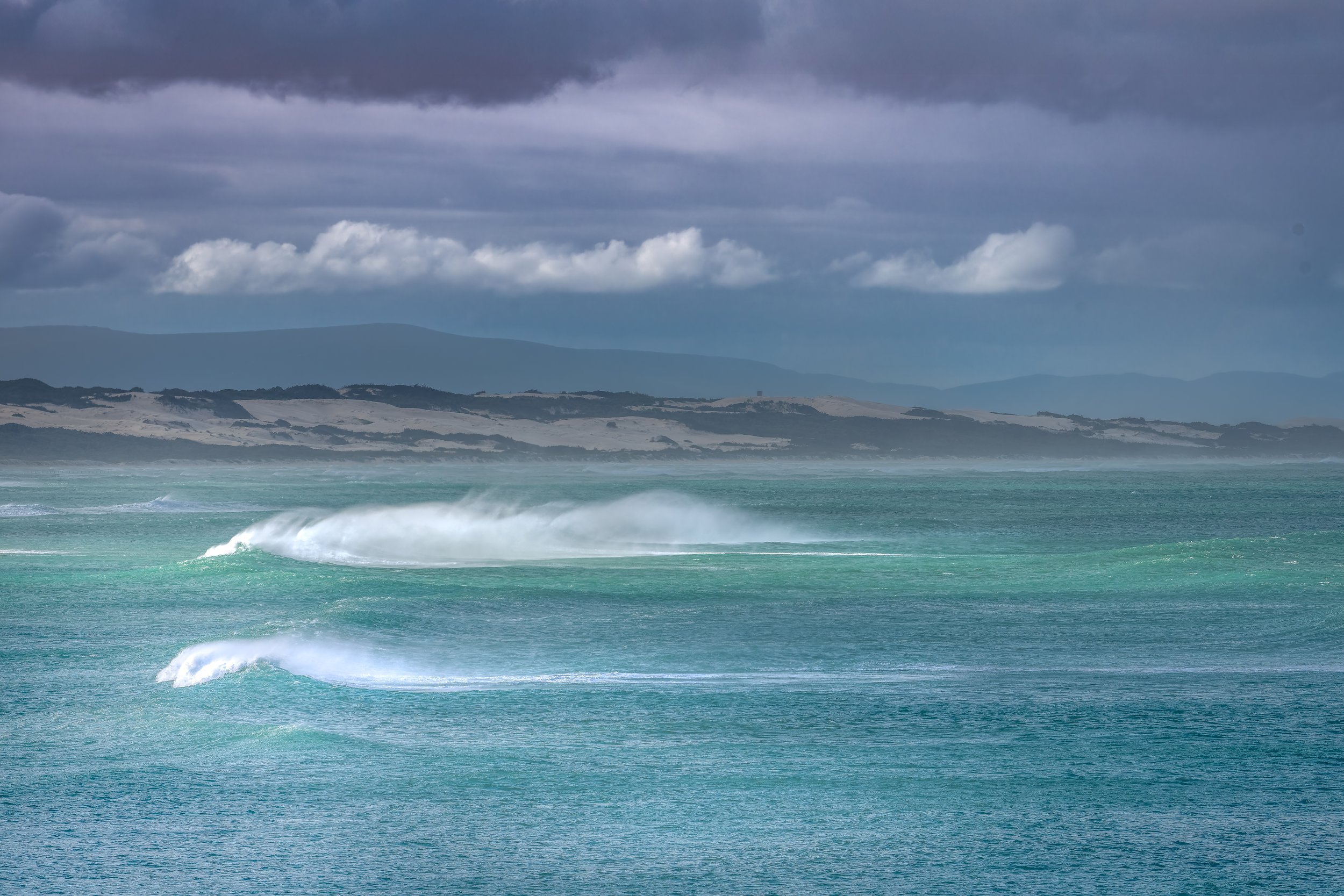 Overcast sky over a choppy ocean with distant landforms and mountains.