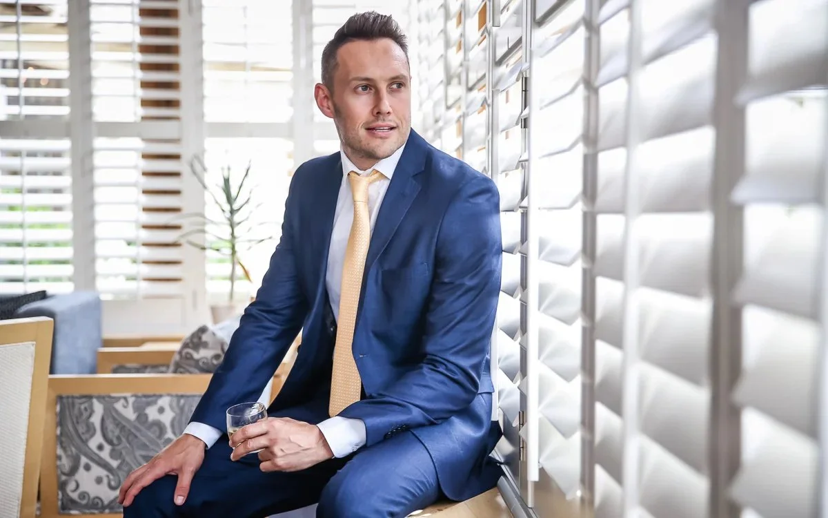 Man in blue suit sitting by window blinds holding a glass