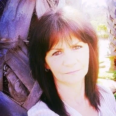 Woman with dark hair posing outdoors near a tree, with bright sunlight illuminating the background.