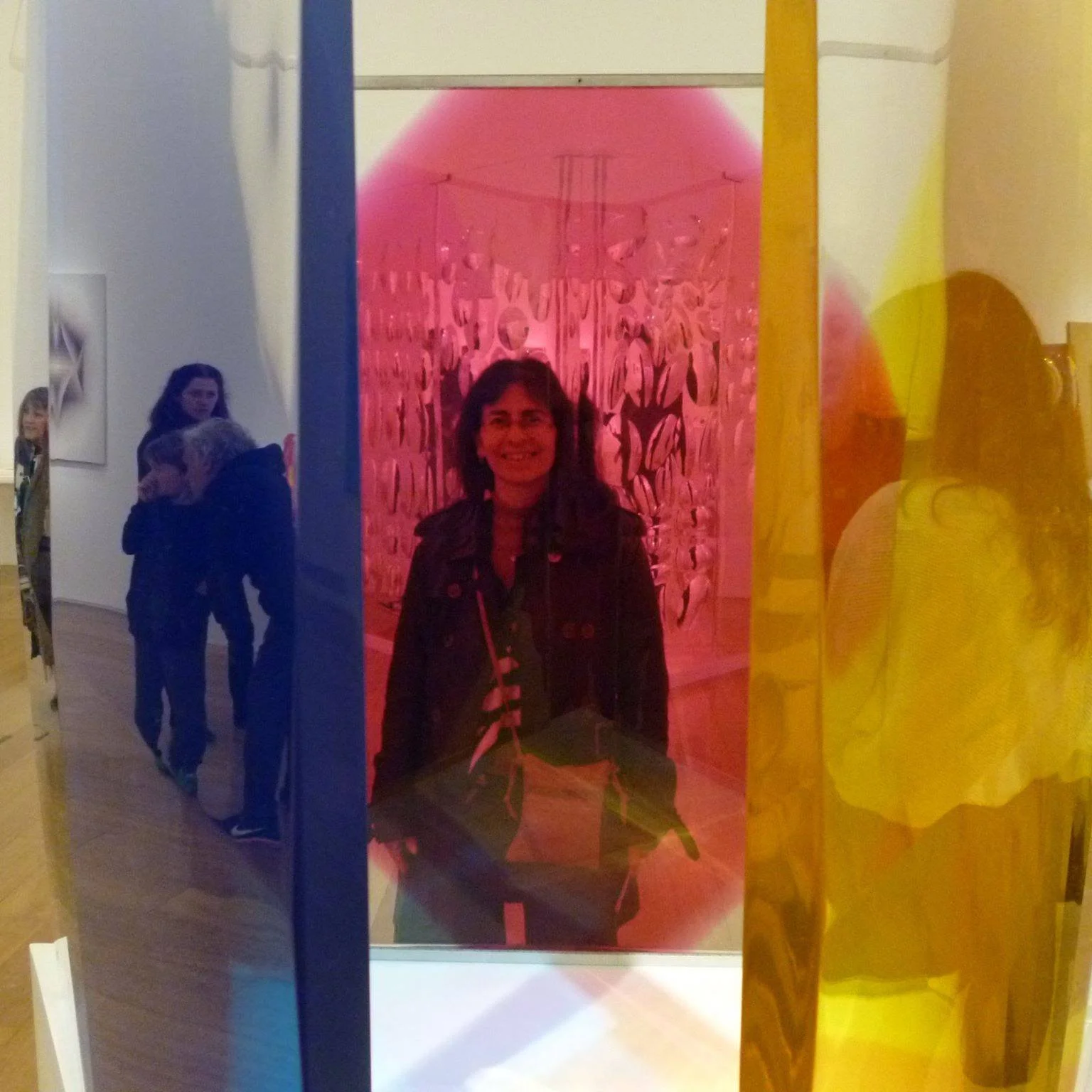 Gallery with colorful transparent panels, with a person visible through the red panel, and reflections of visitors on the blue and yellow panels.