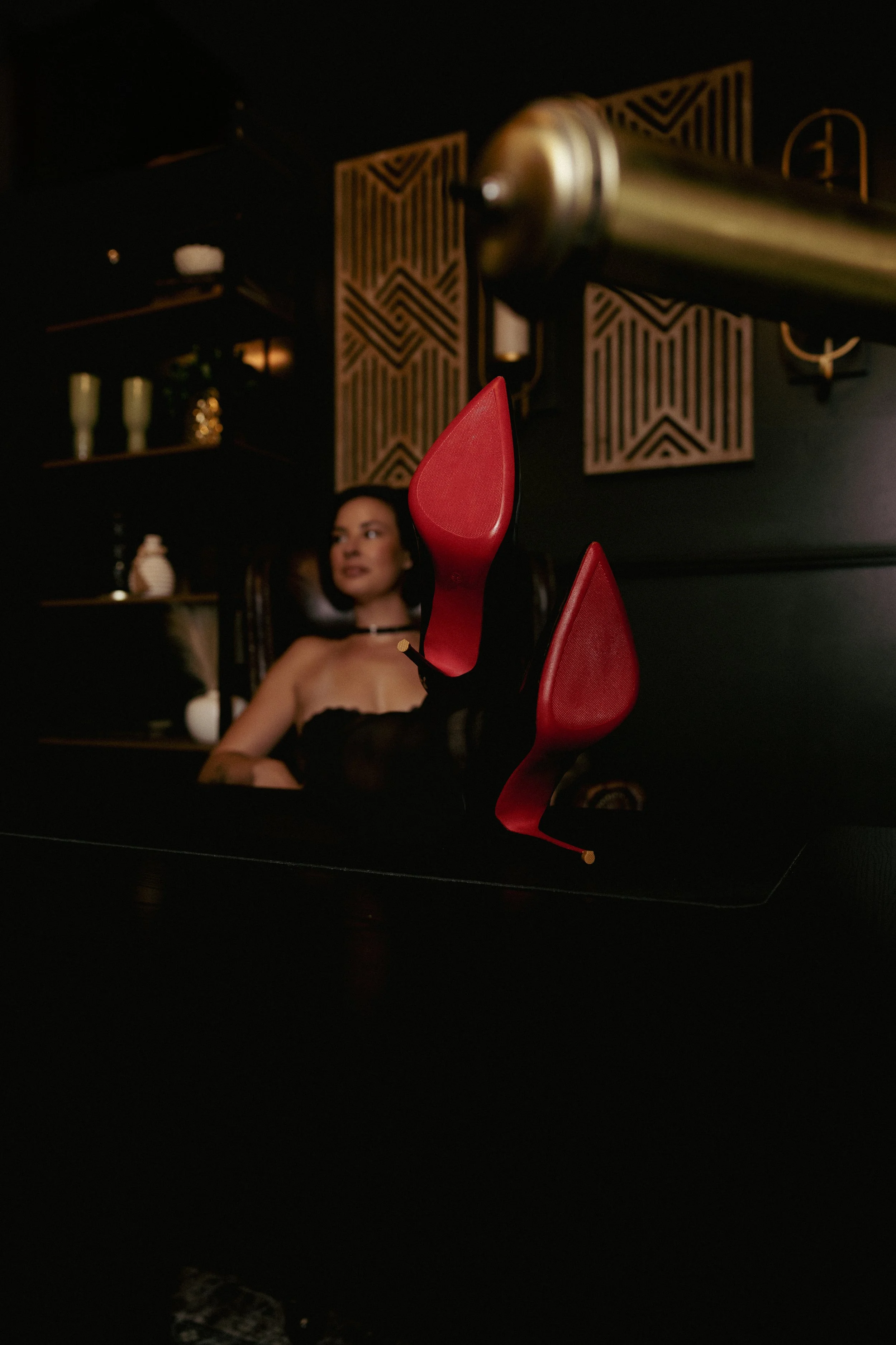 Brunette woman in a boudoir-style office setting with red-bottomed high heels propped on the desk, creating a confident, playful pose.