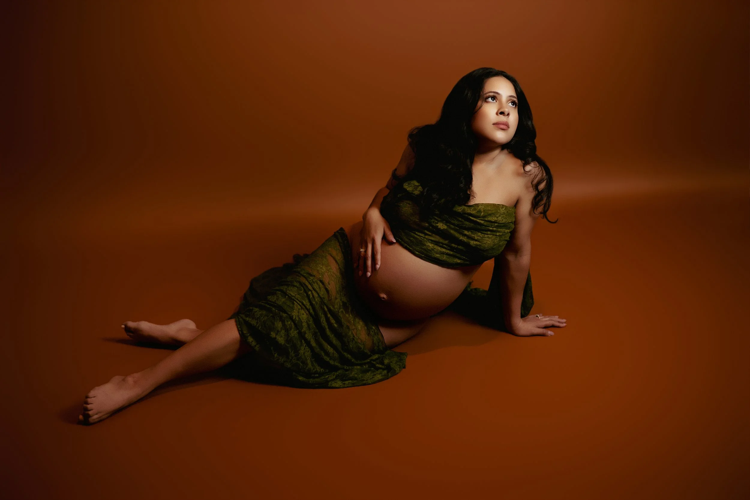 Pregnant woman lying in front of a brown seamless backdrop, draped in green lace with her belly exposed in a maternity boudoir pose.