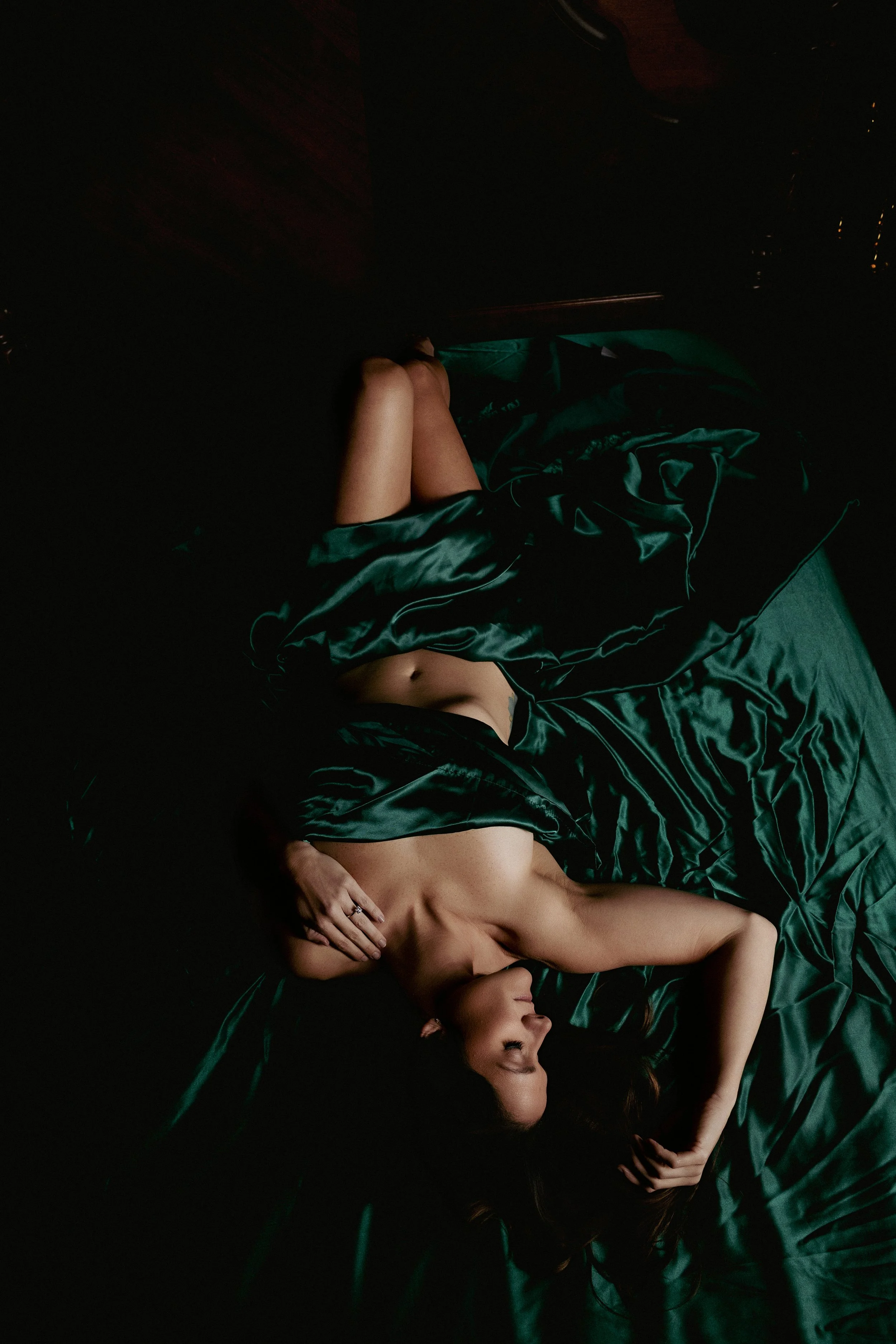 A woman lying on a bed, wrapped in a dark green satin sheet with her head resting on her hand, in a dimly lit room.