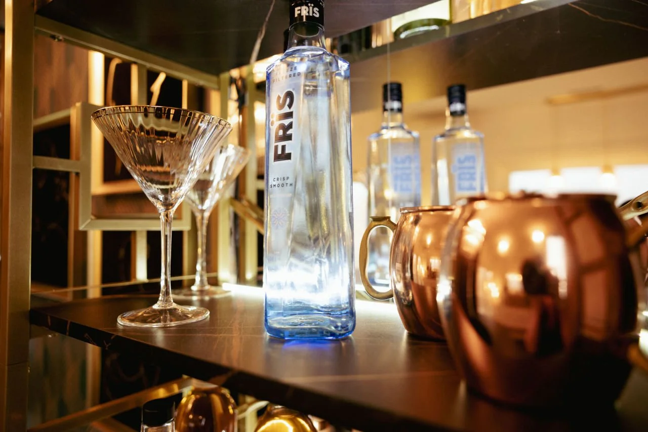 A close-up of a bar shelf displaying a clear bottle of FIRES vodka, two martini glasses, and a copper teapot, with more bottles of FIRES vodka visible in the background.