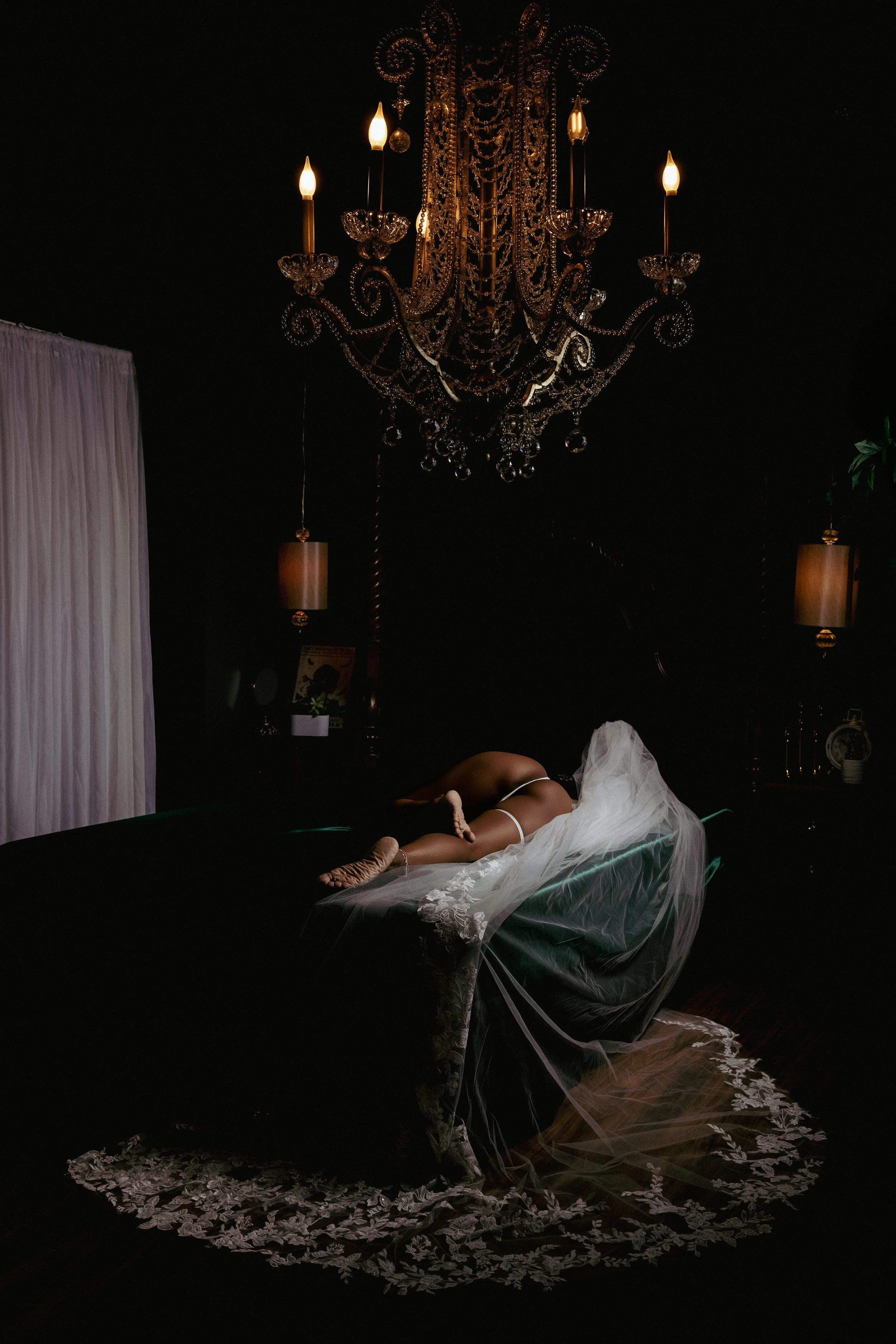 A bride reclines beneath a grand chandelier during a luxury bridal boudoir session at Bondage & Bows Boudoir Photography in Clayton, NC. Moody lighting, flowing veil, and elegant styling create a timeless portrait that celebrates confidence, beauty, 