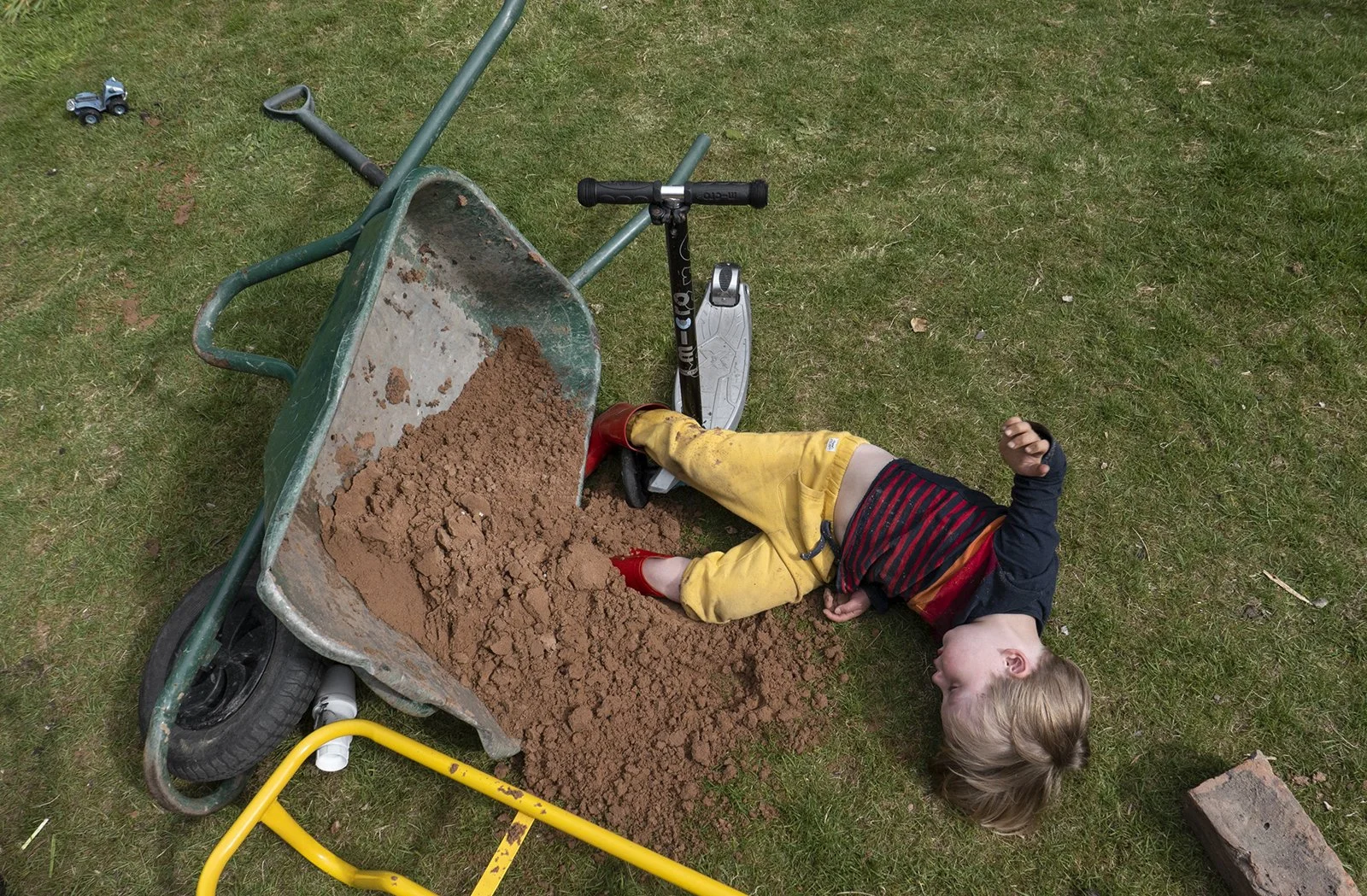Child lying on grass next to overturned wheelbarrow filled with dirt and a scooter