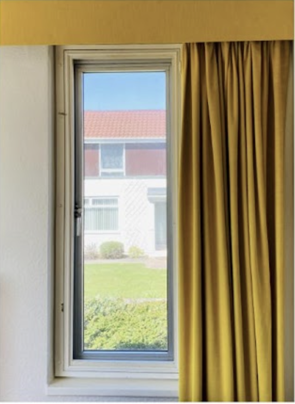 View through a window with yellow curtains partially open, showing an outdoor scene with a building and greenery.