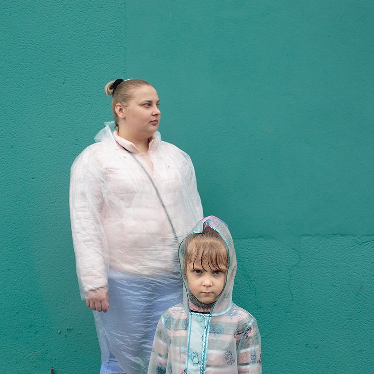 A woman and a child wearing transparent rain ponchos stand in front of a teal wall. The child has a hood on and appears slightly wet.