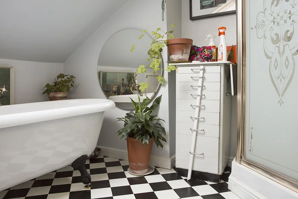 Bathroom with clawfoot tub, checkered floor, plants, round mirror, storage cabinet, and cleaning supplies.