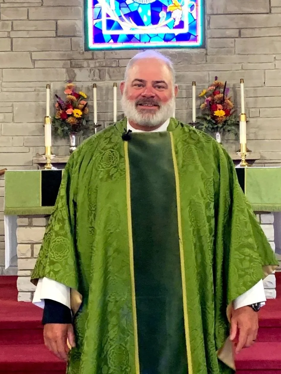 A man standing in front of an altar inside a church, wearing green liturgical vestments, with a brick wall, candles, flower arrangements, and a stained glass window in the background.