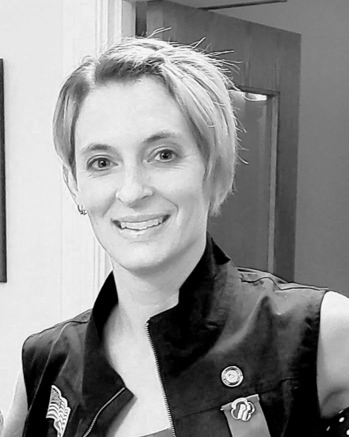 Black and white photo of a woman with short, styled hair, smiling at the camera. She is wearing a sleeveless jacket with various pins and patches, standing indoors with a door in the background.