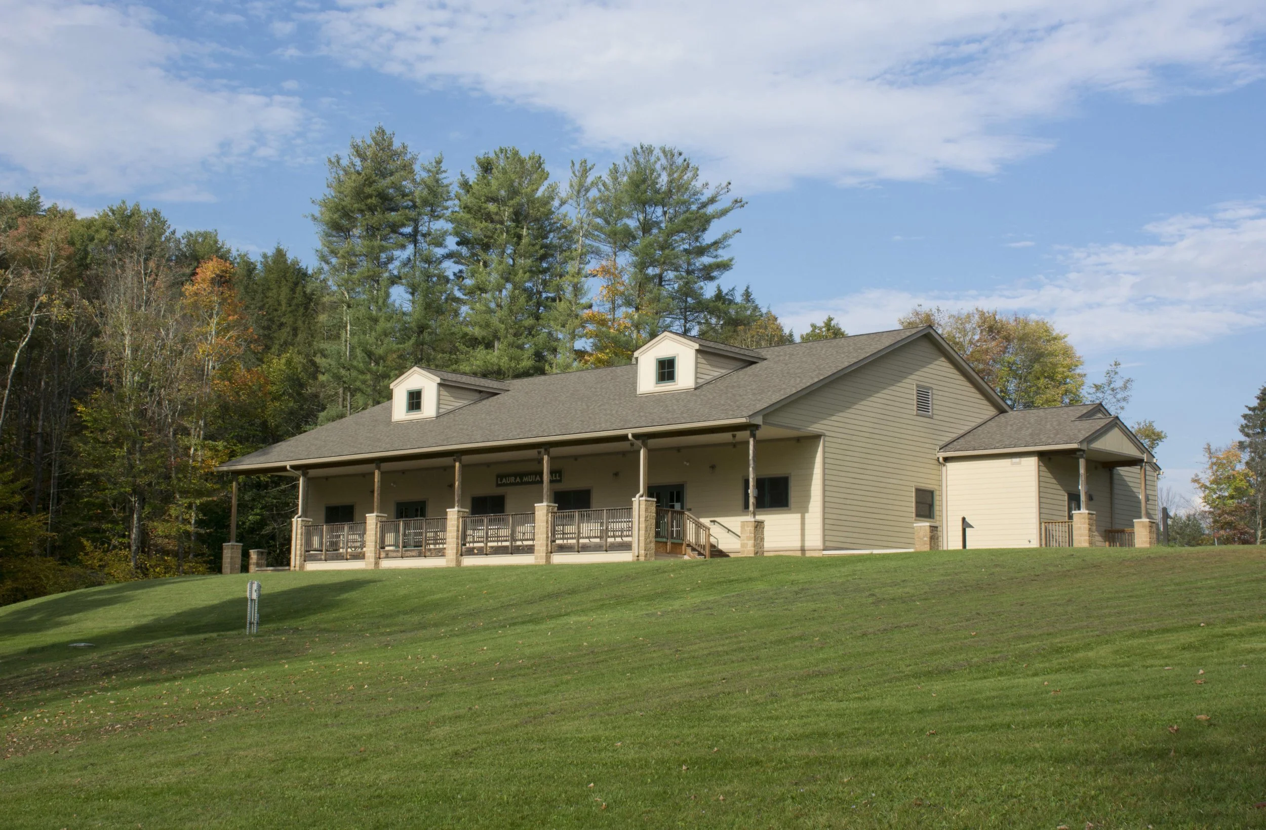 A large house with a beige exterior, a porch with wooden railings, and a sign that reads 'Laura Muir Hall'. It is set on a grassy hill with trees in the background and a partly cloudy sky above.