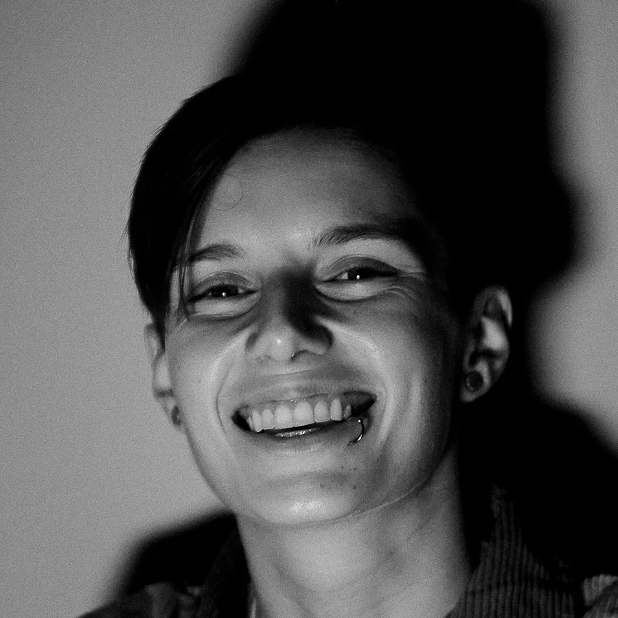Black and white close-up of a smiling person with short hair, pierced lip, and earrings, looking directly at the camera.